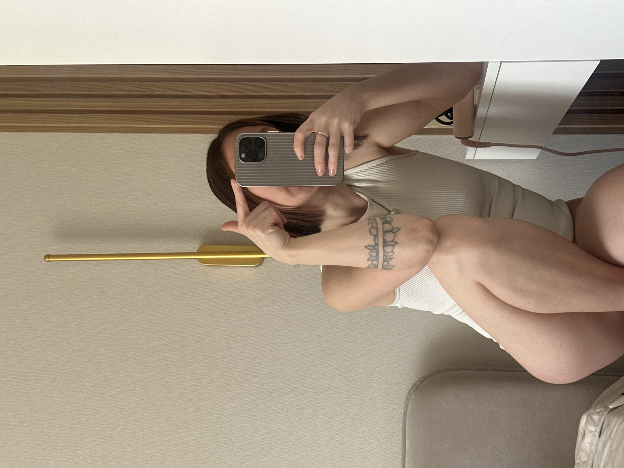 A woman takes a selfie in a mirror. She is seated with her legs crossed. She is wearing a light-colored tank top and has a tattoo on her left arm. She is giving the middle finger with her left hand while holding a smartphone in her right hand to take the picture. There is a beige wall with a gold fixture to the left and a white desk with a power cord plugged into it to the right. The location cannot be determined from the image, but per the prompt, it is assumed to be Votkinsk, Russia.