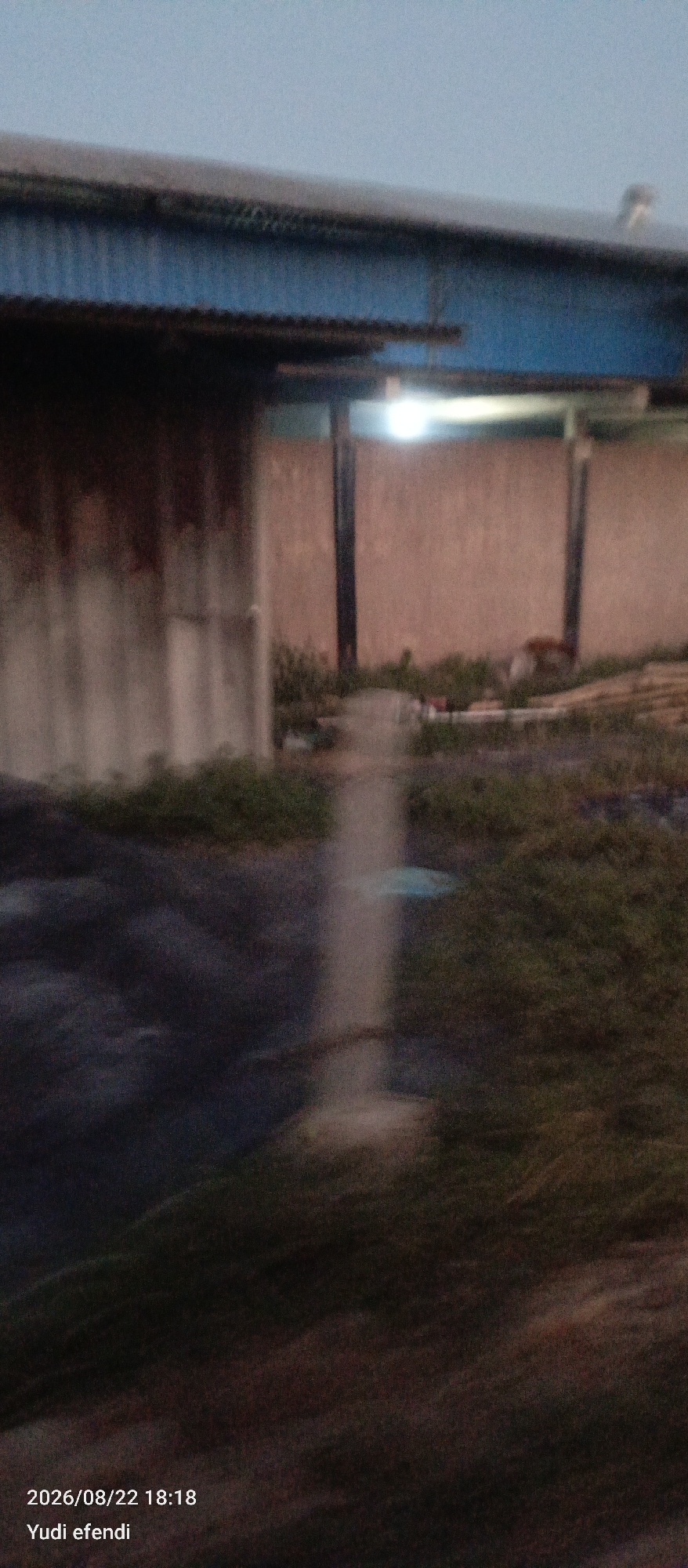 A blurred, low-light image captures an outdoor scene with an industrial or warehouse-like structure dominating the upper portion. A blue corrugated metal roof overhangs a wall made of corrugated metal sheets that appear to be stained with rust or dirt. In the background, there are tan-colored walls with two dark vertical supports, suggesting an open-air structure or an extension of the building. A bright light source, possibly a lamp, is visible in the upper central area. The ground level is covered in vegetation, appearing as a mix of grass and darker, possibly muddy or tarpaulin-covered areas. A faint, vertical light streak extends from the light source down towards the ground, indicating motion blur or a light artifact. The bottom left of the image contains text indicating the date "2026/08/22 18:18" and the name "Yudi efendi". The overall impression is of an industrial or storage area in Rawajitu Selatan, Indonesia, captured in low light conditions.