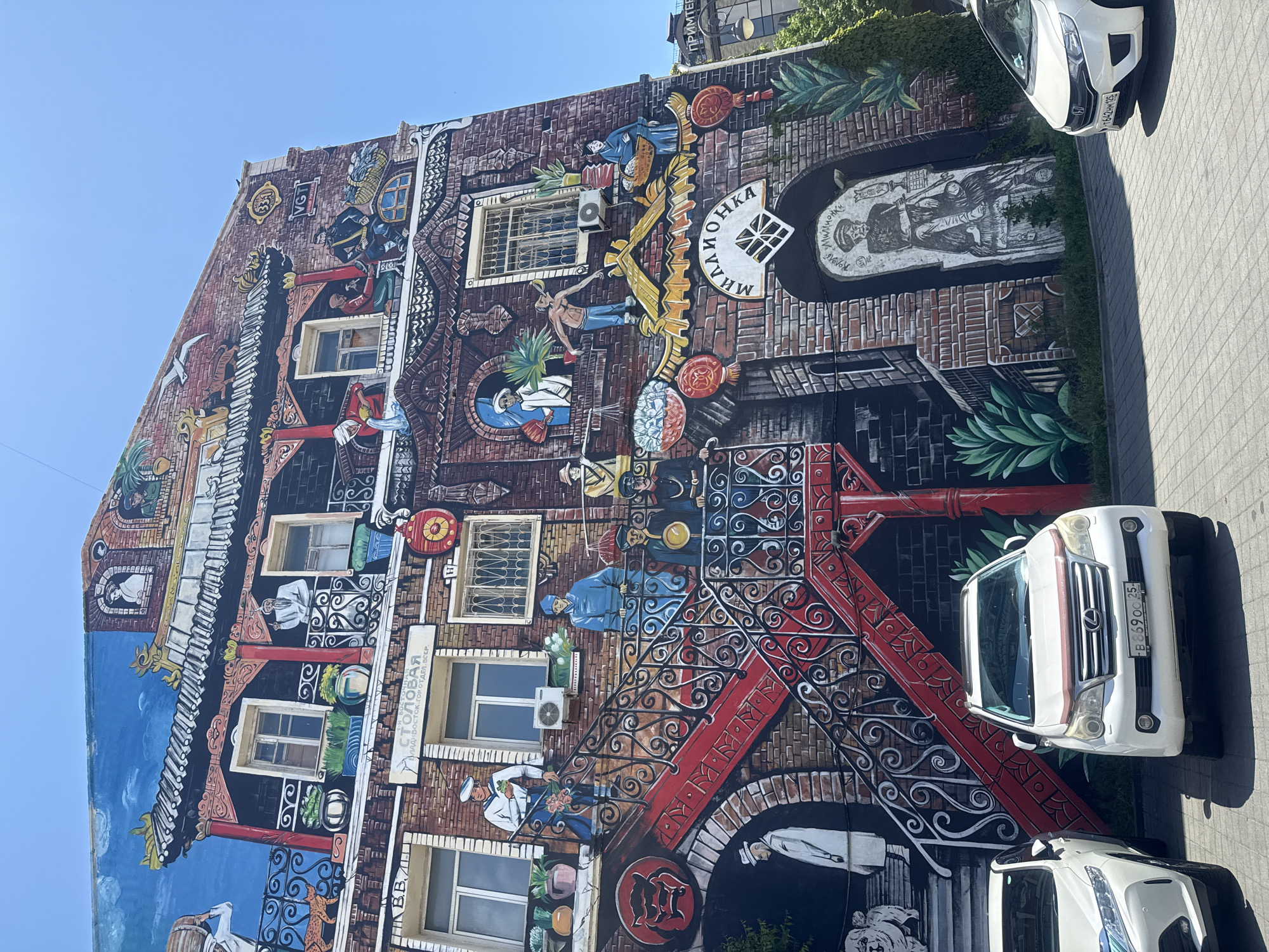 The image shows a large mural on the side of a building in Vladivostok, Russia, depicting various scenes and figures, possibly representing historical or cultural elements of the city. The mural features brickwork, windows, stairs, balconies, and numerous painted figures in traditional attire. There are also some modern elements present, such as air conditioning units on the building and cars parked in front.
Specifically, on the mural, we see:
* A prominent red staircase with ornate black railings.
* Figures on balconies and by windows, some interacting, others seemingly observing.
* A sign above a window that appears to read "СТОЛОВАЯ" which translates to "Canteen" or "Dining Hall".
* A sign with Cyrillic text that appears to read "МИЛЛИОНКА" which refers to Millionka, an old Chinese quarter in Vladivostok.
* A large arched niche on the lower part of the mural containing a black and white drawing of a seated figure.
* Chinese architectural elements, such as curved roofs and decorative patterns.
* Potted plants and foliage integrated into the mural.
In the foreground, three cars are visible:
* On the left, a white car, partially visible.
* In the center, a white Lexus GX SUV with a license plate that appears to be B 869 OO 25.
* On the right, a white Honda Fit with a license plate that reads Y 764 HH 125.
The environment appears to be a sunny day with a clear blue sky. The ground in front of the building is paved with interlocking bricks. Greenery is visible on the right side of the image, with ivy climbing up a wall and some trees. The overall impression is a vibrant and detailed piece of street art adorning an urban building.