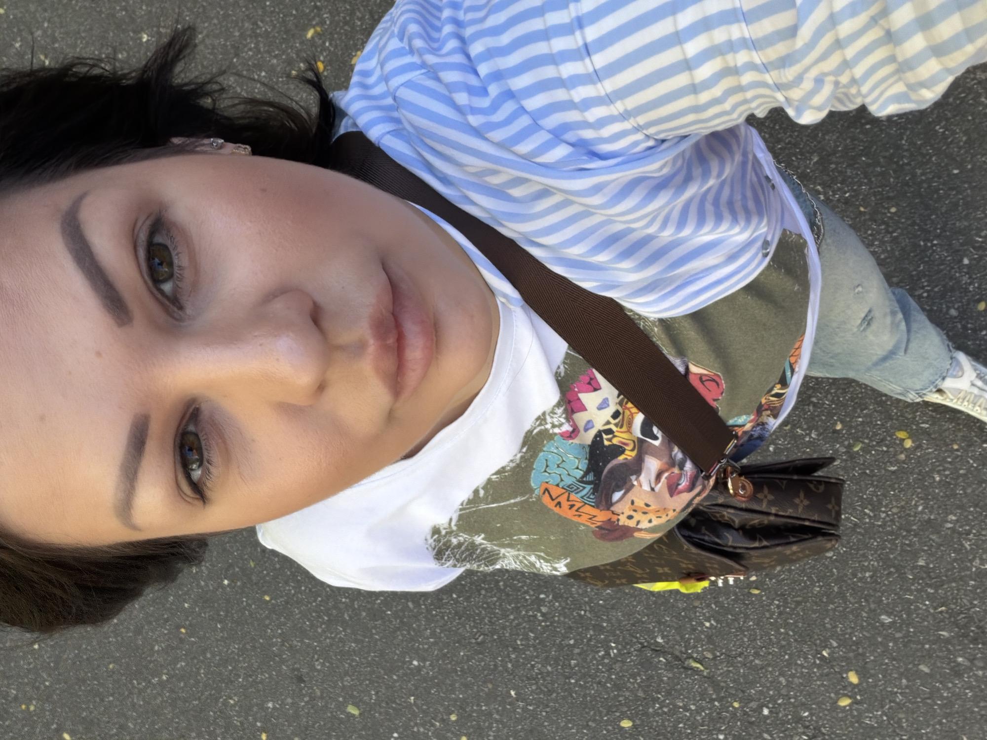 The image depicts an adult female positioned at an elevated angle, taking a selfie outdoors in the city of Sochi, Russia. She has dark brown hair, brown eyes, and is looking directly at the camera with a neutral expression. Her face shows light makeup, including defined eyebrows and minimal eye makeup. She is wearing two small earrings on her visible right ear, one a small hoop and the other a stud. Her attire consists of a white crew-neck t-shirt layered under an olive green t-shirt featuring a vibrant, multi-colored graphic print of stylized faces or heads with varied patterns, including leopard print and geometric designs. Over these, she wears a light blue and white vertically striped long-sleeved button-down shirt, partially open, with sleeves pushed up. She is also wearing light blue denim jeans with a distressed detail on the left knee, and white sneakers are partially visible. A dark brown cross-body bag, consistent with a Louis Vuitton Monogram pattern, with gold-toned hardware and a solid brown strap, is worn across her body. A silver-colored chain or bracelet is visible on her left wrist. The ground is dark asphalt with scattered small yellow leaves or debris, indicating an outdoor, daytime setting.