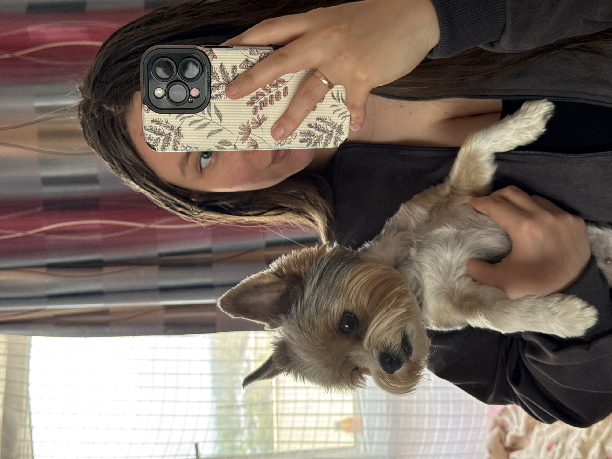 A woman is holding a small, light brown and white terrier dog in her arms, while taking a selfie with her mobile phone. The woman's face is partially obscured by her phone, which has a light-colored case with a floral design. She has brown hair and is wearing a dark-colored jacket. The background appears to be a room interior with window blinds. There is no information to indicate the location in the city of Krasnodar, Russia.