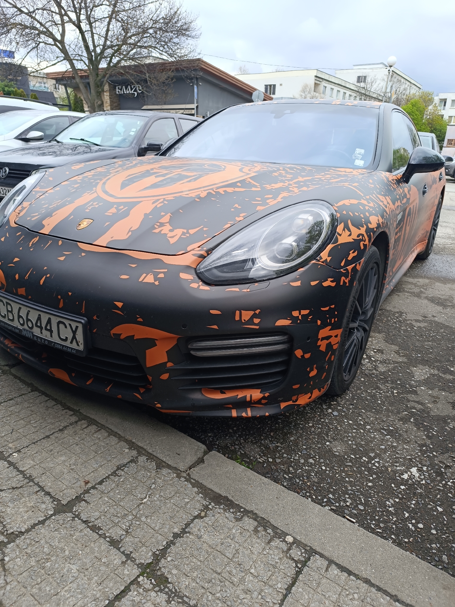 The image depicts a matte black Porsche Panamera with an orange, stylized splatter pattern parked on a city street. The car has a Bulgarian license plate that reads "CB 6644 CX." The street is paved with a mix of concrete slabs and asphalt. In the background, other cars are parked, along with buildings and trees under an overcast sky. A restaurant with a sign written in Cyrillic, "БЛАГО," is also visible. The location is Sofia, Bulgaria.