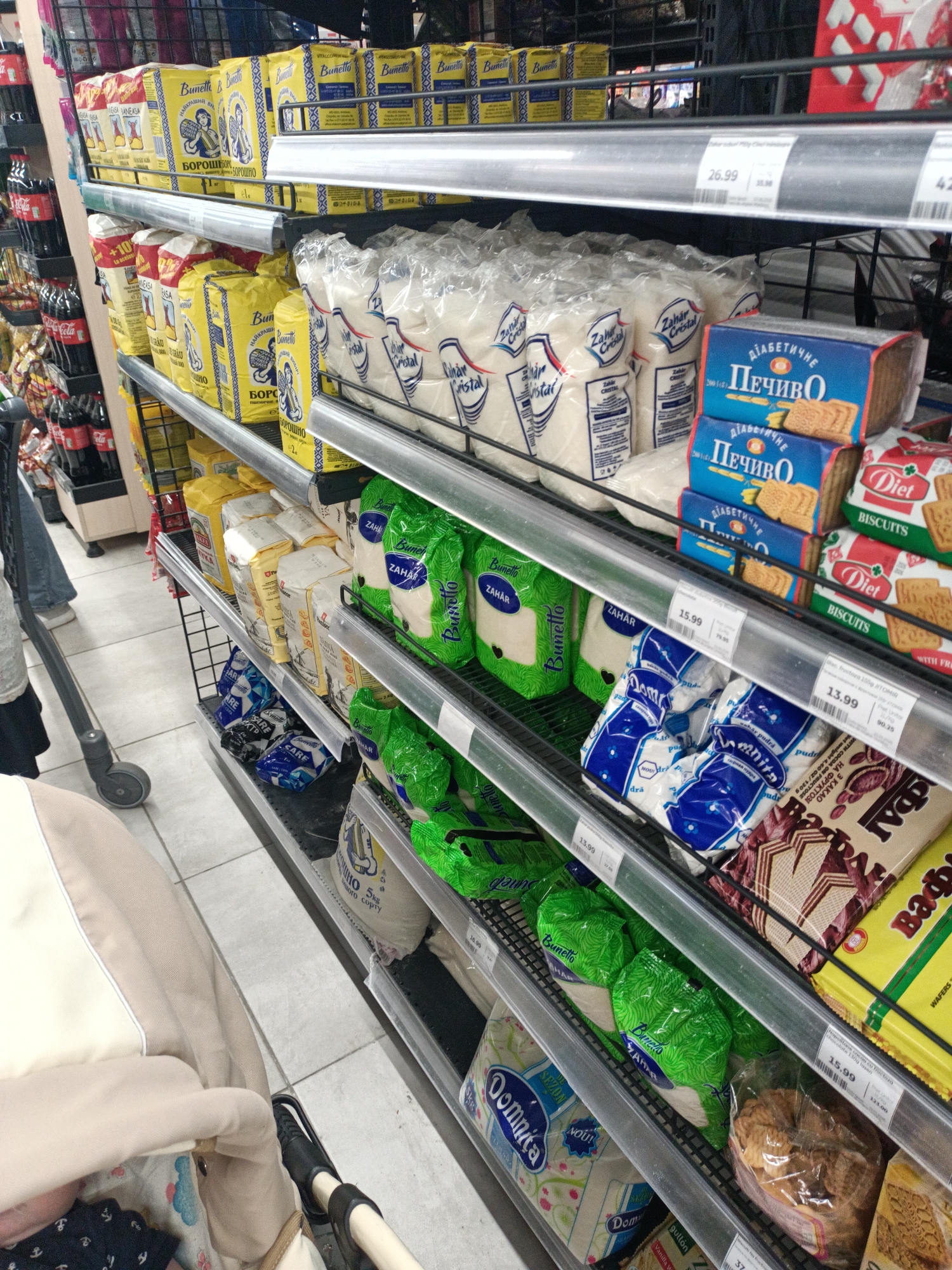 The image shows a grocery store aisle with shelves stocked with various food products. The shelves are made of metal racks. On the top shelves, there are boxes of "Bunetto" brand sugar and "Pecibo" brand biscuits. Below these, there are several bags of white sugar labeled "Zahar Cristal". Further down, there are packages of flour and other food items. A shopping cart with a baby inside is partially visible on the left side of the image. The floor is tiled. The lighting appears to be artificial, typical of an indoor retail environment.