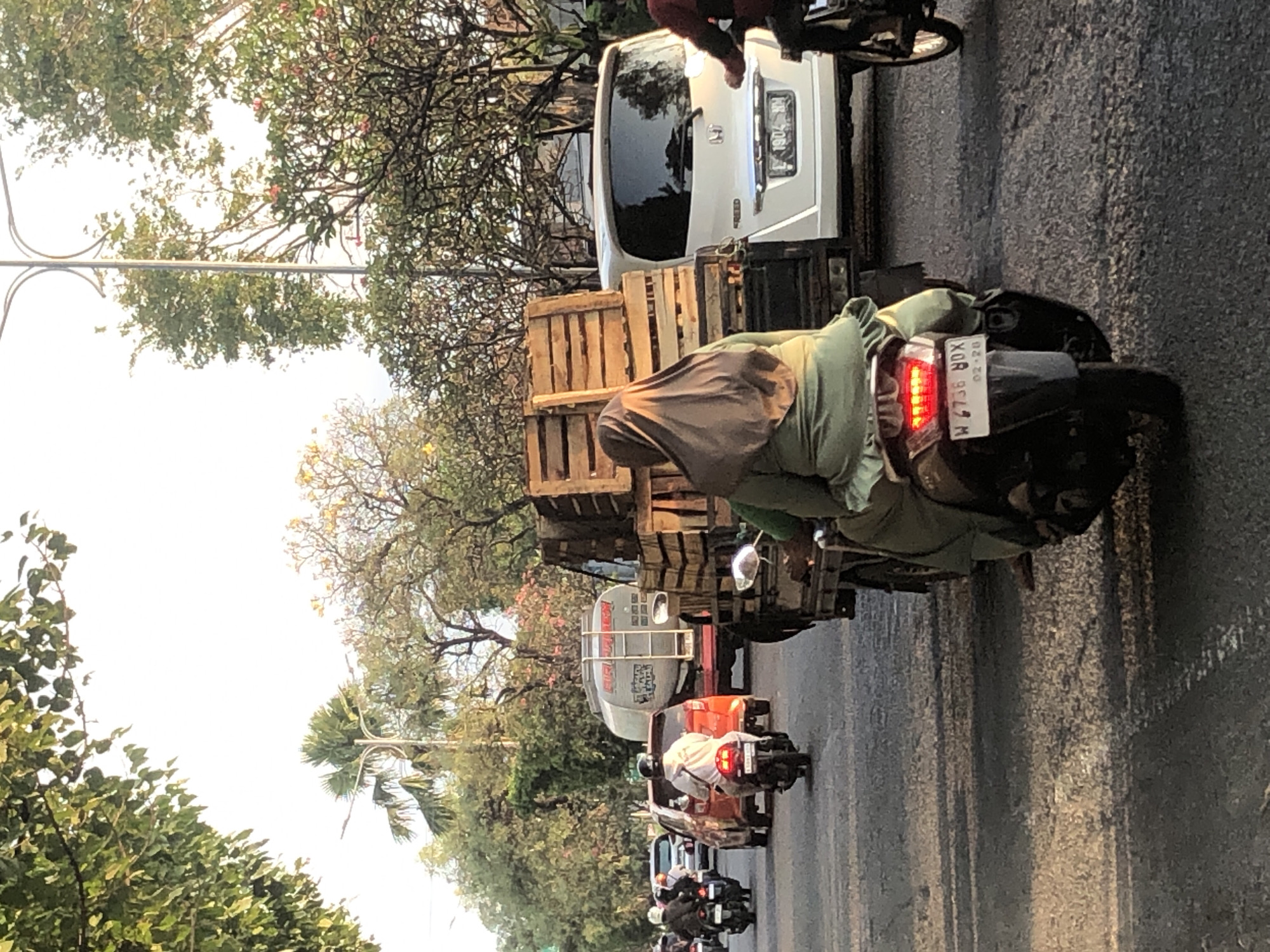 The image depicts a street scene in Gelam, Indonesia, featuring a traffic jam with several vehicles and individuals. In the foreground, a person is riding a scooter, wearing a green outfit and a brown hijab. The scooter has a license plate with the characters "W 4736 BDX". Behind this scooter, a truck is visible, loaded with wooden crates. To the right, a white car, possibly a Honda, is parked, with its license plate reading "L 1904 NM". Further back, other vehicles, including a red car and a tanker truck, are part of the traffic. Trees line the sides of the road, and the sky appears bright, suggesting daytime. The overall scene captures a moment of congested urban traffic in a tropical setting.
