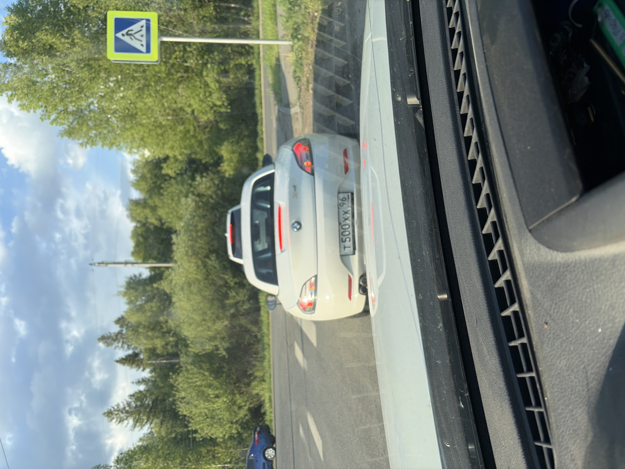 The image shows a white BMW car with a Russian license plate T 500 XX 96, partially visible, on a road in Perm, Russia. A pedestrian crossing sign is visible near the car. The photo is taken from inside another vehicle. The weather appears sunny with some clouds in the sky. Trees line the roadside.
