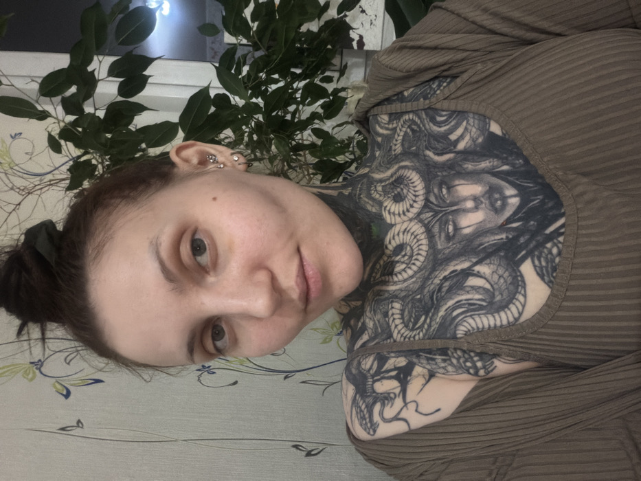 A fair-skinned woman with brown hair pulled back into a bun poses indoors. She wears a brown, ribbed top. Extensive black and grey tattoos cover her chest and upper shoulder, depicting Medusa. She has multiple piercings on her left ear. A potted green plant stands behind her. The background features a light-colored wall with a floral pattern. The image is rotated slightly counter-clockwise.