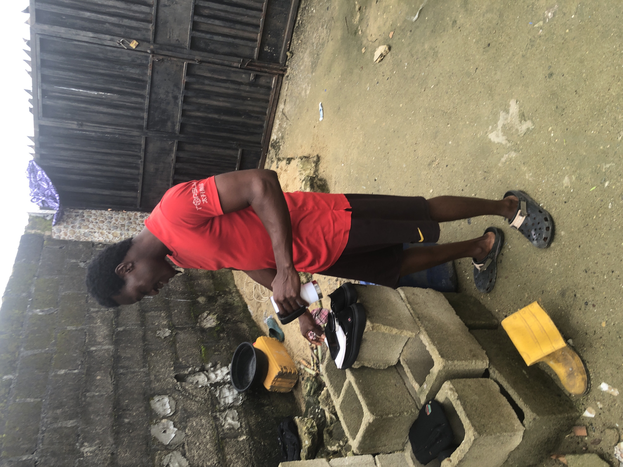 In Ado, Nigeria, a man stands in an outdoor, somewhat unkempt area next to a gray cinder block wall. He is wearing a red t-shirt and dark shorts, and is holding a white object, possibly a cleaning device or small appliance. In front of him, on a stack of cinder blocks, are several pairs of shoes, including black and white sneakers. To his left, a yellow rubber boot lies on the ground. Behind him, a dark metal gate is visible, along with a purple patterned cloth draped over the wall. The ground is a mix of concrete and sand. The overall impression is of a utilitarian, outdoor workspace.