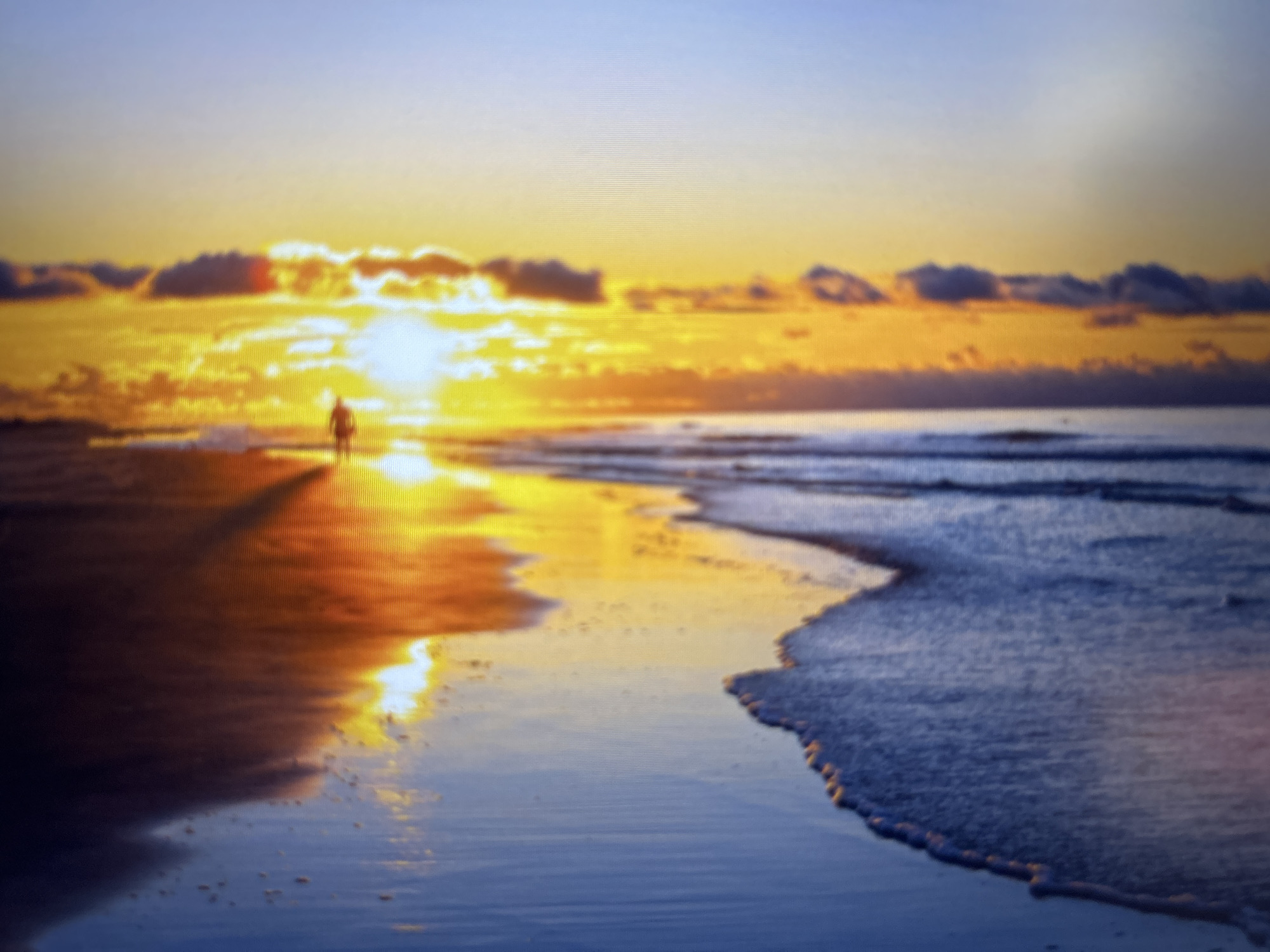 A blurred photograph depicts a beach scene at sunset. The sun is low on the horizon, shining brightly through a layer of clouds. A silhouetted figure is walking on the sand towards the sun. The wet sand reflects the sunlight, creating a mirror-like effect. Waves are gently lapping the shoreline. The location is likely a beach in Varna, Bulgaria, given the prompt's context.