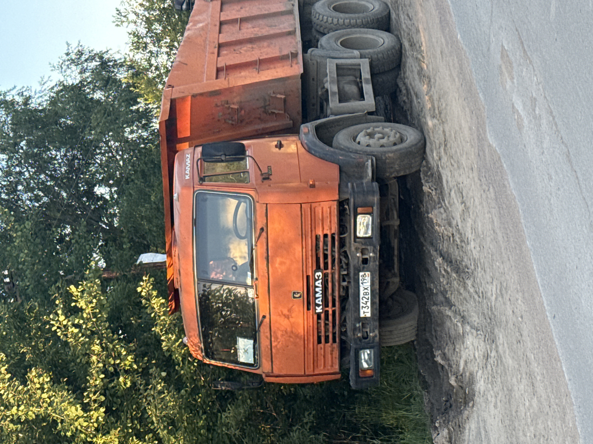 An orange Kamaz dump truck is parked on a dirt road next to a tree line. The truck's license plate reads "T342BX 198". The truck appears to be in a rural or industrial area outside of Saint Petersburg, Russia. There is a person in the driver's seat of the truck, but their face is not visible. The truck's bed is empty. The ground is covered in mud and tire tracks. The sky is clear and blue.