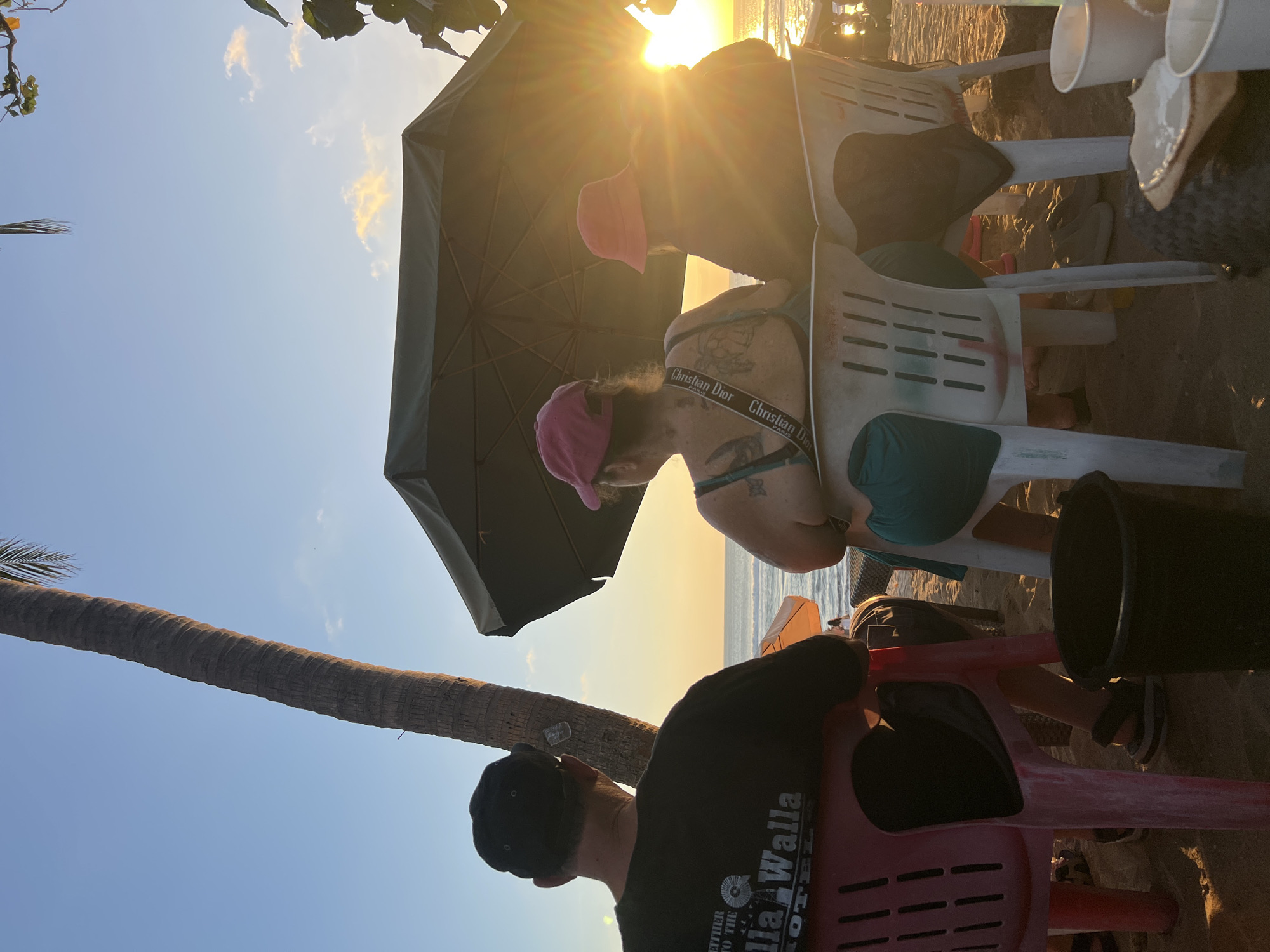 In Kuta, Indonesia, three individuals are seated on chairs on a beach, facing the ocean and the setting sun. A man in a black t-shirt and baseball cap is seated on the left. To his right, a woman with a pink hat and teal top is seated. Another person, also wearing a pink hat, is seated to the right of the woman. A large umbrella provides shade, and the sun is visible behind it, casting a warm glow. Palm trees and vegetation frame the scene. The ground is covered in sand, and various objects, including cups and a bucket, are present. The water is calm, with gentle waves lapping at the shore.