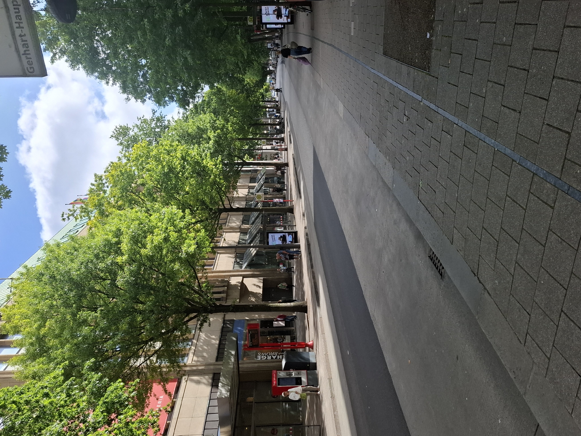 The image depicts a wide, tree-lined street in Hamburg, Germany, likely during daytime on a sunny day with some clouds. The street is paved with asphalt and has a pedestrian sidewalk made of gray interlocking pavers on the right side. Buildings with modern architecture, featuring light-colored facades and large windows, line the left side of the street. Many lush green trees with full canopies are planted along the sidewalk, casting shadows on the street.
Several people are visible on the sidewalk. On the left side, a woman in a light-colored hat and jacket stands near a red and gray kiosk-like structure. Further down the sidewalk, a few individuals are walking and standing, some near shop entrances. On the right side of the street, a woman with dark hair and a dark top is walking away from the camera, carrying shopping bags. Further down the sidewalk, two people are standing and conversing near a bus stop shelter with a digital advertisement.
Prominent signage includes a street name sign at the top right that reads "Gerhart-Haupt". There are also various commercial signs on the buildings and advertisements on display. The overall impression is a busy urban environment with a strong emphasis on pedestrian access and greenery.