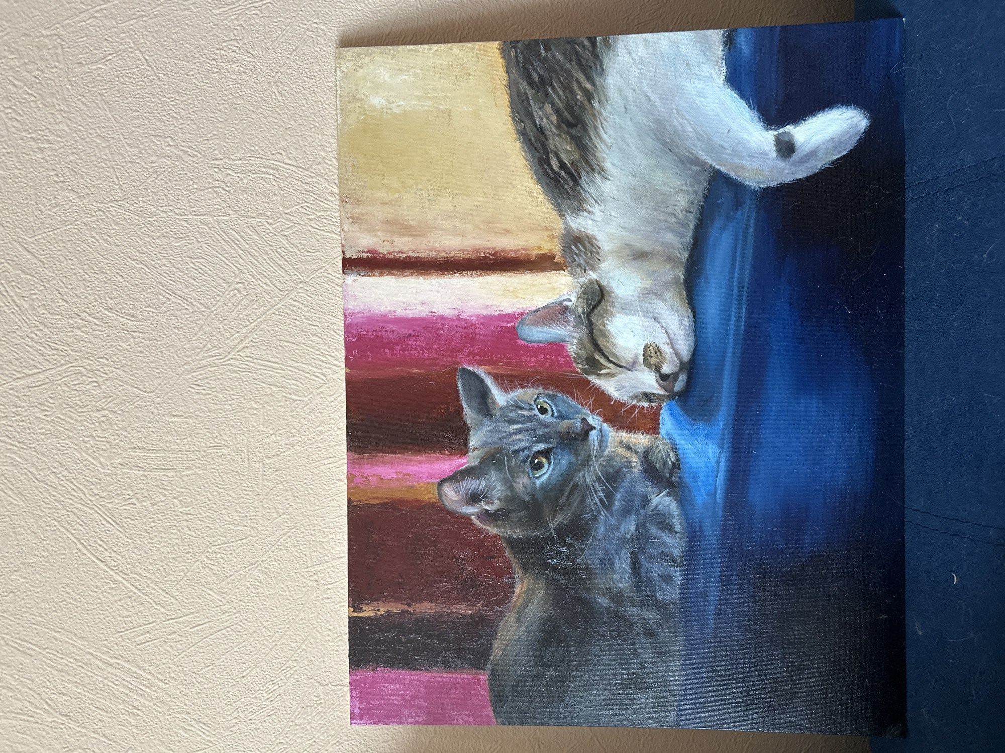 The image depicts an oil painting on canvas featuring two cats. The canvas is leaning against a wall with beige textured wallpaper. To the right of the canvas is a blue surface.
One cat is positioned upside down, its head near the center of the canvas, while the other cat looks up at it. The upside-down cat has white and grey fur, while the other cat is grey. The background of the painting includes horizontal bands of yellow, brown, pink, and dark red. The right side of the painting features blue.
The painting is not located in Krasnoyarsk, Russia.