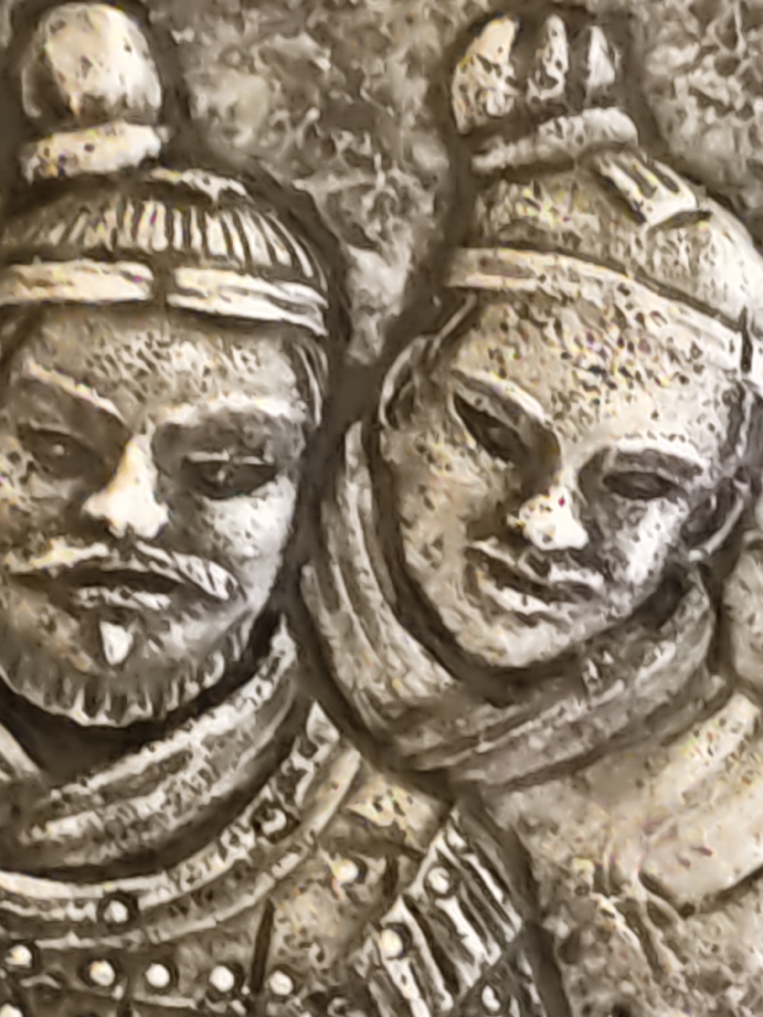 This image is a close-up, low-resolution photograph of a bas-relief sculpture depicting two figures in what appears to be ancient or historical attire. The sculpture is made of a rough, textured material, likely stone or plaster, with a light, weathered appearance.
The figure on the left has a stern expression, with visible facial features including a mustache and goatee. They are wearing a helmet with decorative bands. The figure on the right has a softer facial expression and is also wearing a helmet. Both figures appear to be wearing layered clothing or armor, with distinct patterns and embellishments visible on their chests and necks. The background of the sculpture is textured and indistinct, suggesting it is part of a larger work or a wall.
There is no information in the image or the provided context to suggest this sculpture is located in Rostov-on-Don, Russia. The description is based solely on the visual elements of the photograph.