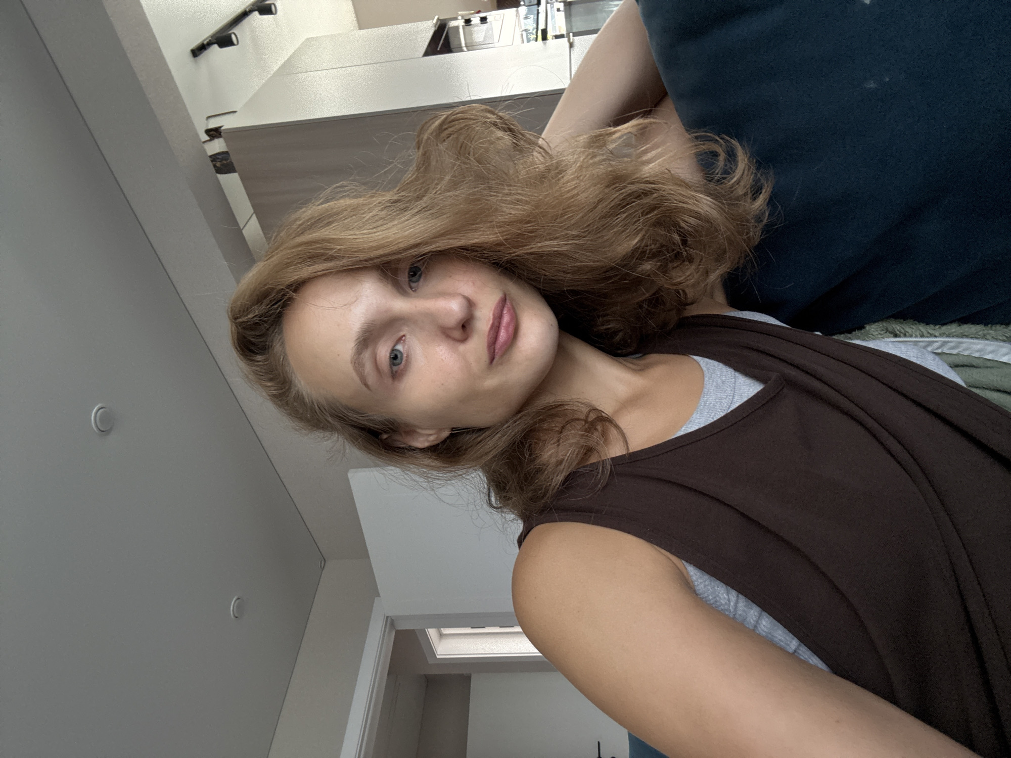 A young woman with light brown hair is captured in a close-up, indoor selfie. She is wearing a brown tank top over a grey camisole. Her left arm is resting on a dark blue surface, possibly a couch. The background includes a ceiling with recessed lighting fixtures, a white door, and kitchen cabinetry. The image is taken from a slightly elevated angle, looking down at the woman. The woman's face is turned slightly towards the camera, with her blue eyes looking directly into the lens. Her expression is neutral, with her lips slightly parted. The overall lighting is soft and natural, suggesting daylight. There is no indication of the specific location within the city of Подшивалово, Russia, as the image contains only generic indoor elements.