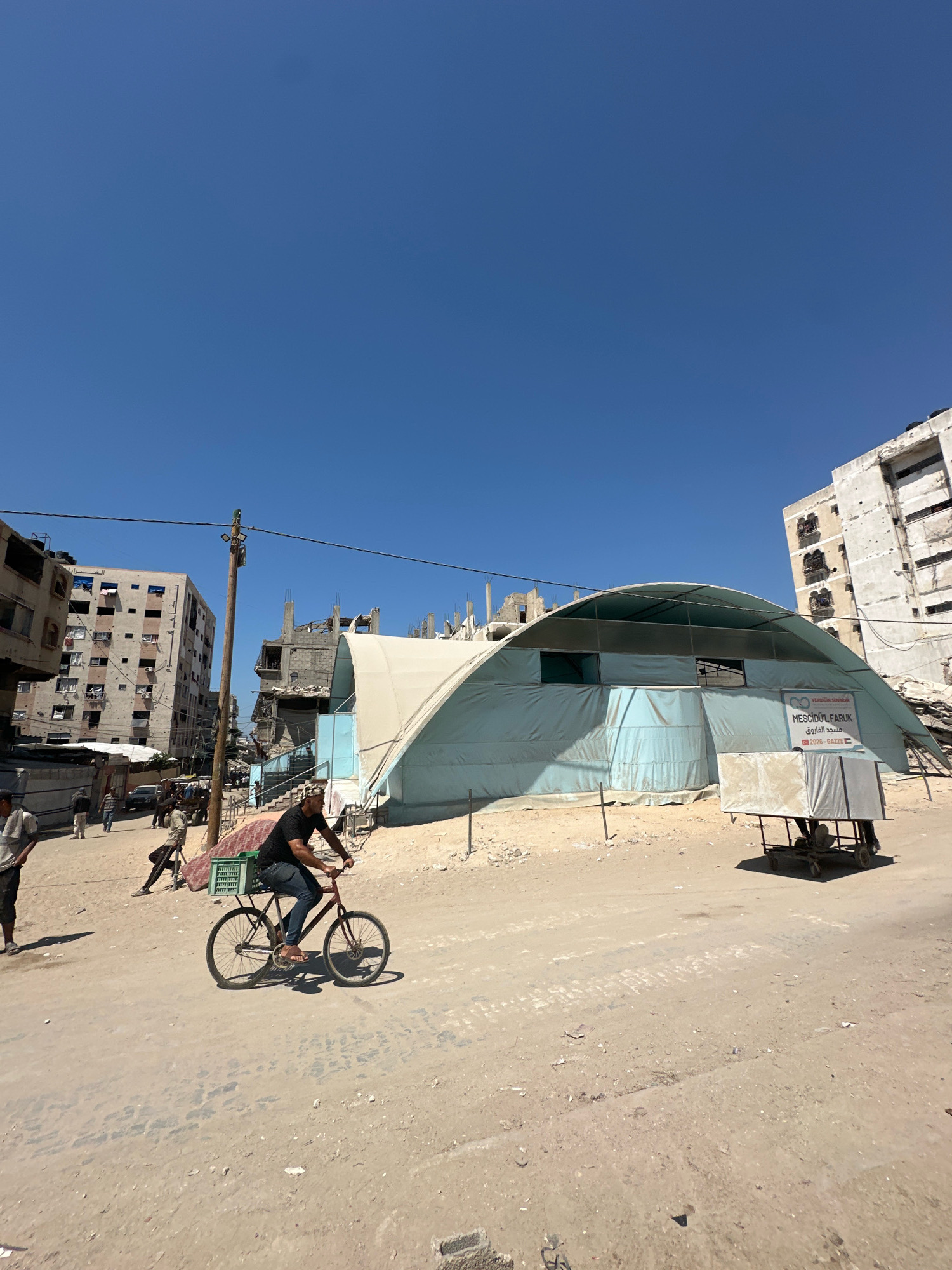 In the city of Gaza, Palestinian Territories, a clear blue sky presides over a dusty street scene. The street is lined with damaged buildings, some appearing recently bombed out, with exposed rebar and crumbling concrete. In the foreground, a man cycles on a bicycle, a green crate strapped to the back. Behind him, a large, light blue tent structure with a curved roof is erected, likely a temporary shelter or community space. To the right of the tent, a cart covered with a light-colored tarp is parked. In the background, more damaged buildings and construction elements suggest ongoing reconstruction or further destruction. Several other people are visible in the scene, walking or standing near the buildings. The overall impression is one of devastation and resilience in a war-torn urban environment.