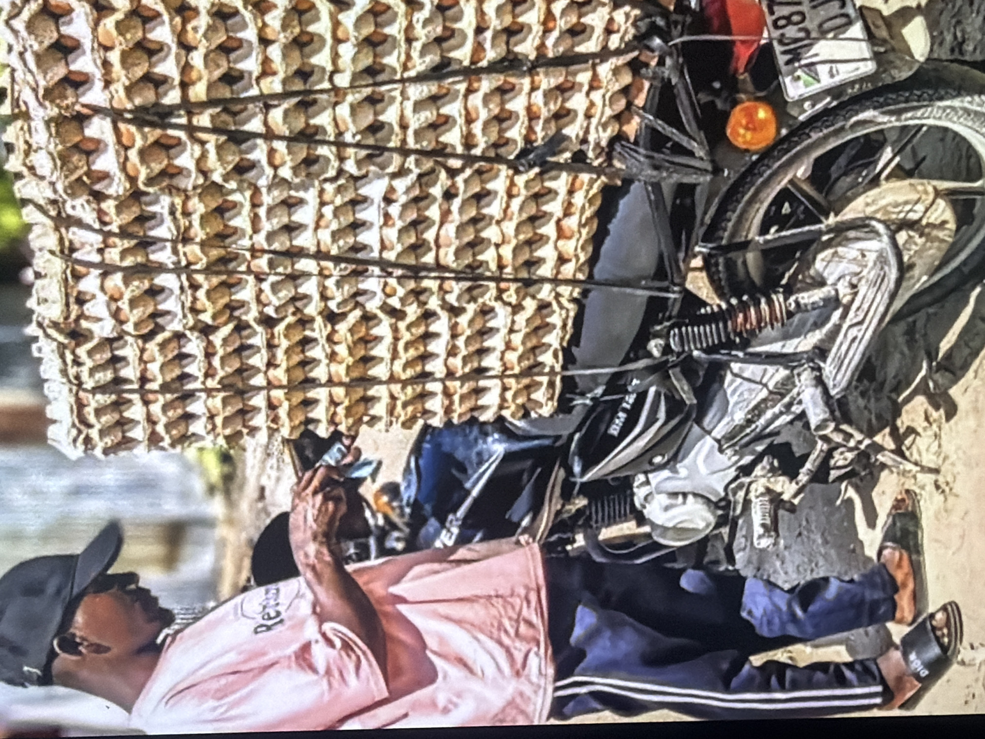 In the city of Bukuru, Nigeria, a man is shown on a dirt road beside a motorcycle laden with stacks of egg cartons. The man, wearing a pink t-shirt and dark pants, is seen holding a small object in his hands near the motorcycle. The motorcycle, a dark color, has a large load of egg cartons strapped to its rear. The cartons are stacked high, creating a towering structure of eggs. The environment appears to be outdoors, with the ground being dirt and the background showing some greenery and a building. The scene is illuminated by natural sunlight.