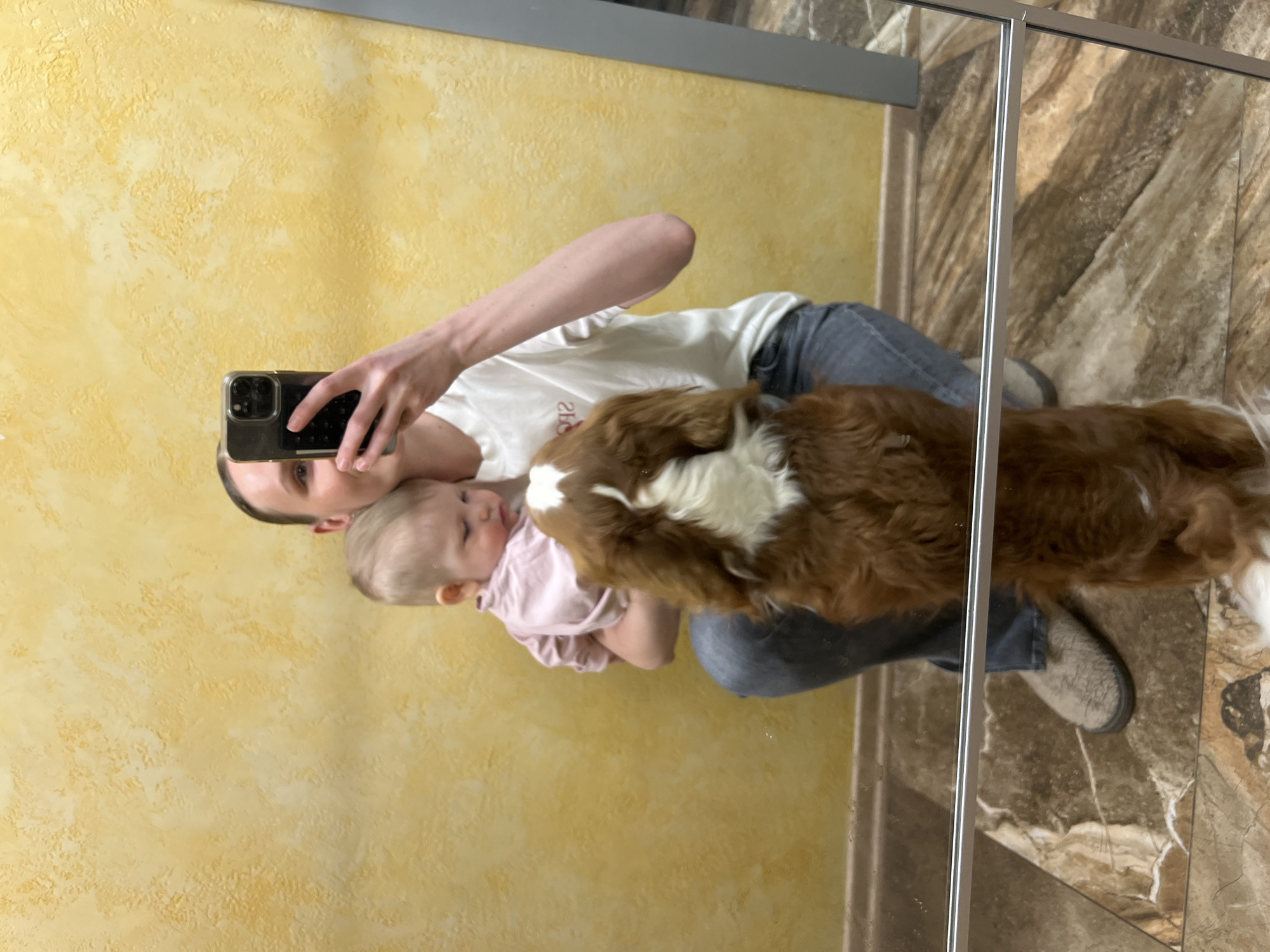 An adult, likely a woman, is taking a mirror selfie indoors in Khimki, Russia. She holds a baby and a medium-sized brown and white dog. The adult, with her head slightly tilted, looks towards her smartphone held up in her right hand to capture the reflection. She is wearing a white t-shirt, on which the reversed text "2U" is visible (meaning "U2" in reality), and blue jeans, with grey furry slippers or shoes on her feet.
The baby, dressed in a light pink top, is cradled in the adult's left arm, looking towards the dog. The dog, with prominent brown and white markings, particularly on its face and chest, is positioned in front of the adult and baby, its head close to the baby. The scene depicts a calm and affectionate family moment.
The background shows a textured, warm yellow wall on the left side of the reflection. On the right, a vertical silver metal frame or divider separates the yellow wall from a section of brown, possibly marbled or tiled, floor and wall. The floor beneath the adult and dog also appears to be a brown marbled or tiled surface. The lighting suggests an indoor setting, but the time of day is not discernible.