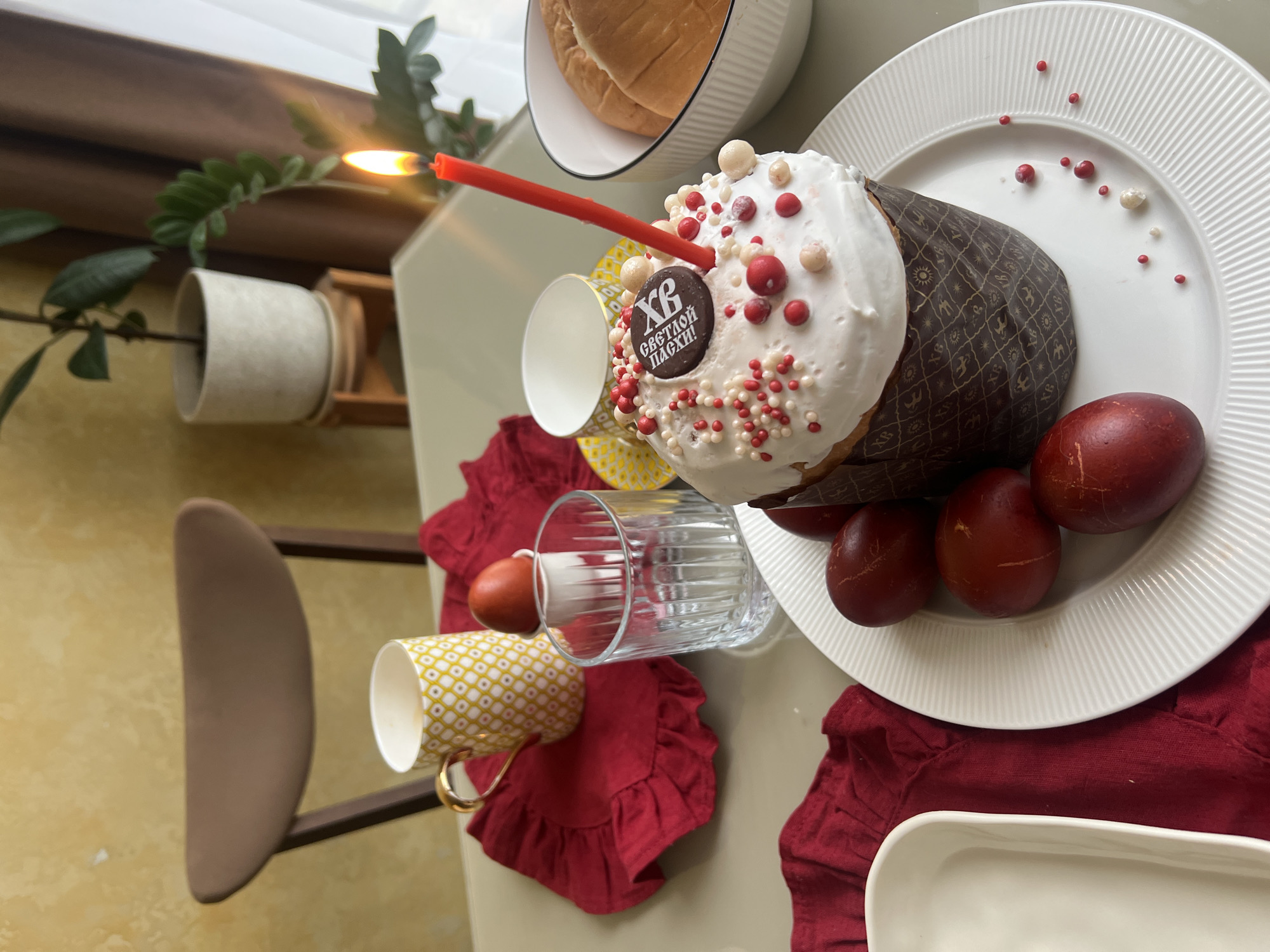 The image depicts a celebratory Easter scene on a table. The central object is a decorated kulich, a traditional Easter bread, adorned with white frosting, colorful sprinkles, and a small chocolate label with Cyrillic text. A lit red candle is inserted into the top of the kulich. Several dyed red Easter eggs are arranged around the kulich on a white plate. Additional objects on the table include a bowl of bread, a patterned teacup, a drinking glass, and red cloth napkins. A houseplant is visible in the background. It's not possible to pinpoint the exact location in the city of Khimki, Russia, from this single image.