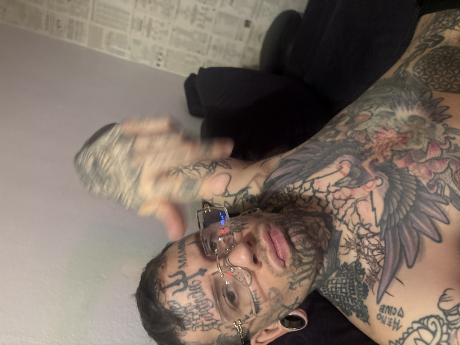 An adult male is depicted in a close-up, angled shot, likely a selfie, in an indoor setting. The individual is heavily tattooed across the face, neck, chest, arms, and hands. Facial tattoos include a trident symbol and "MAMBA" on the forehead, script above the left eyebrow, and various patterns around the eyes, cheeks, and chin. He wears rimless, gold-framed glasses and large ear gauges in both earlobes. His lips are pursed, forming a "duck face" expression. The chest is covered with detailed tattoo work, featuring an eagle or phoenix-like bird, script reading "HERO CLUB," floral designs, and geometric patterns. The right arm is raised and blurred, indicating movement, with the hand out of focus. The background consists of a plain, light-colored wall on the left, transitioning to a wall or surface covered with printed papers or newspaper clippings arranged in a pattern on the right. A dark-colored piece of furniture, possibly a couch or chair back, is partially visible behind the subject on the right. The specific location within Buenos Aires, Argentina, cannot be determined from the visual information provided.