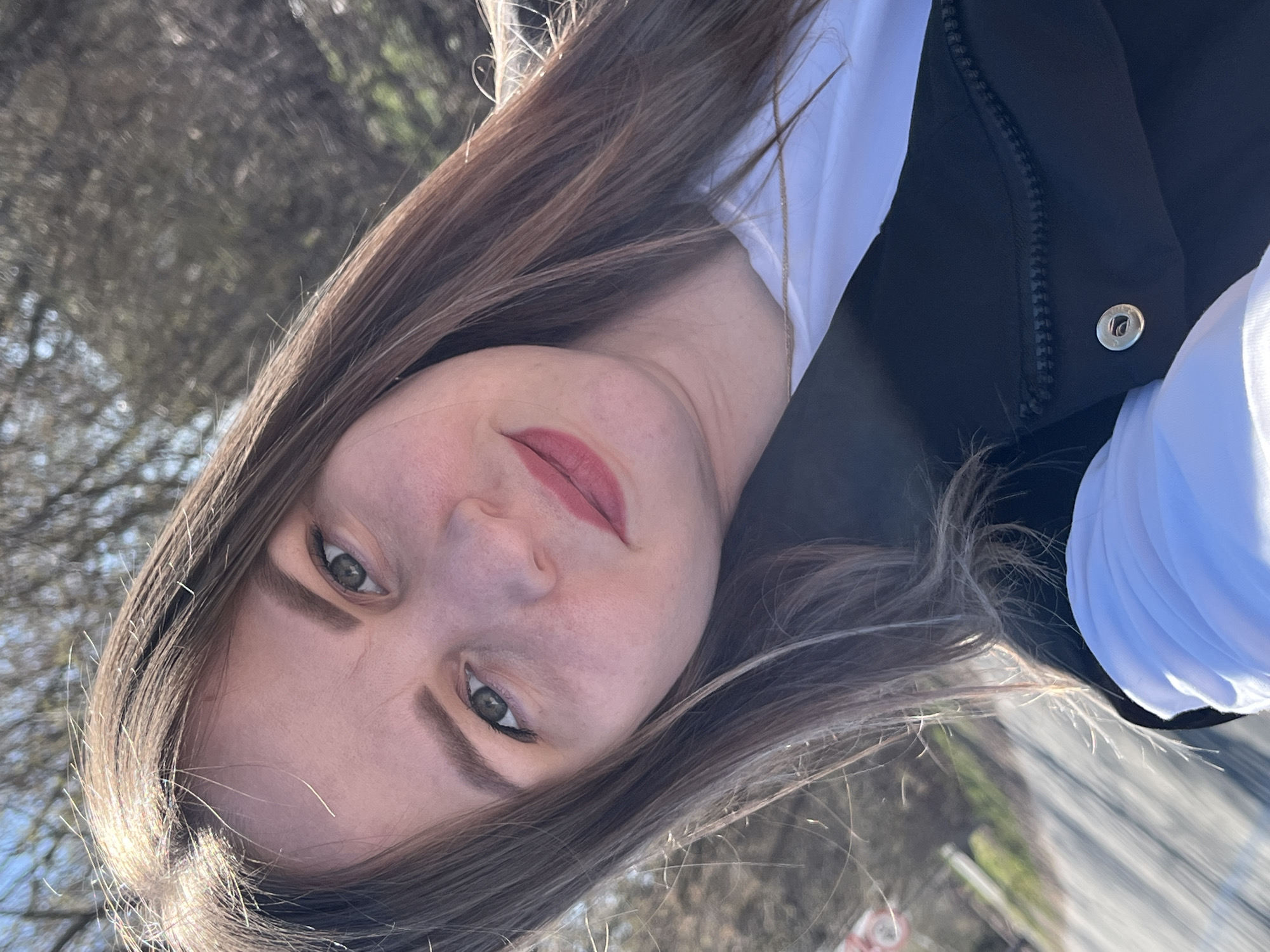A woman with long brown hair is looking directly at the camera. She is wearing a white shirt underneath a black vest. The background appears to be a blurred outdoor environment, possibly near a road or path with trees. The specific location in Zav'yalovo, Russia, cannot be determined from the visual information provided.