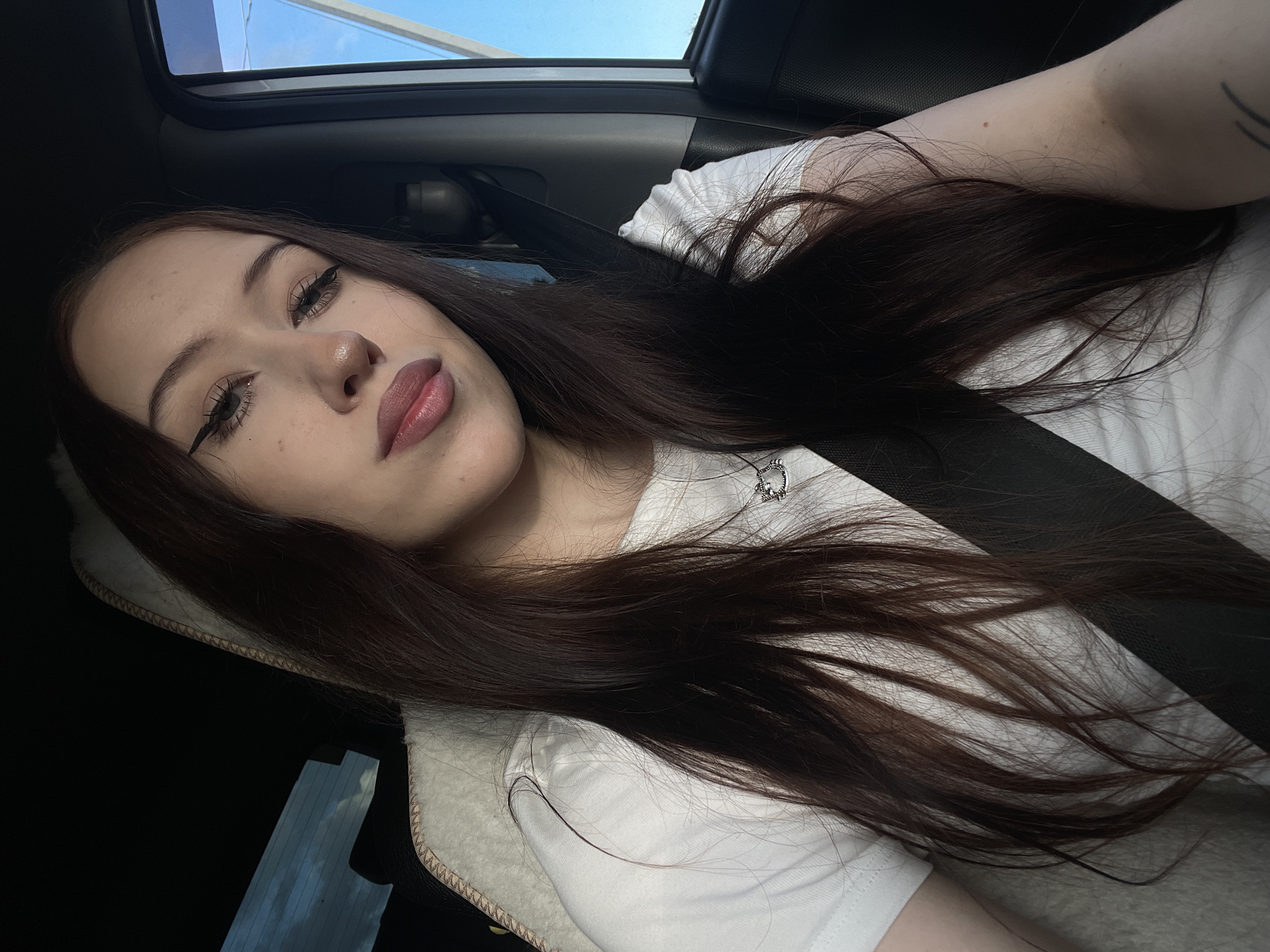 A young woman with long, dark brown hair, wearing a white t-shirt, is seen in a selfie taken inside a car. The car has a sunroof open, revealing a bright blue sky with some clouds, suggesting it is daytime with fair weather. The woman has on winged eyeliner and neutral lipstick. Her hair is spread across her shoulder and chest, and she is wearing a black seatbelt. A small, silver pendant with a 'D' on it is visible on her t-shirt. The scene appears to be a casual moment, possibly during a car ride. The interior of the car is dimly lit, contrasting with the bright sky visible through the sunroof. The image is taken from a slightly elevated angle, looking down at the woman. No text is visible in the image. The location context is Гавриловка, Russia.