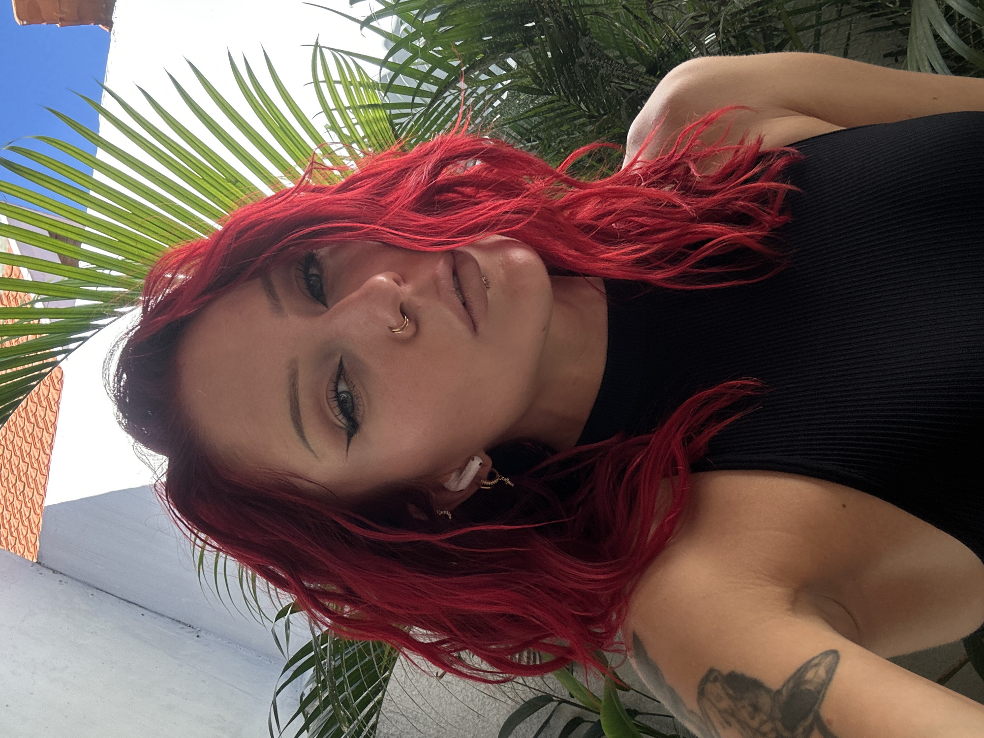 A woman with red hair and fair skin, wearing a black ribbed top, is photographed from a low angle. She has a nose ring and earrings. She has winged eyeliner. Part of a tattoo is visible on her arm. Behind her are palm fronds, a white building, and a blue sky. The location is North Kuta, Indonesia.