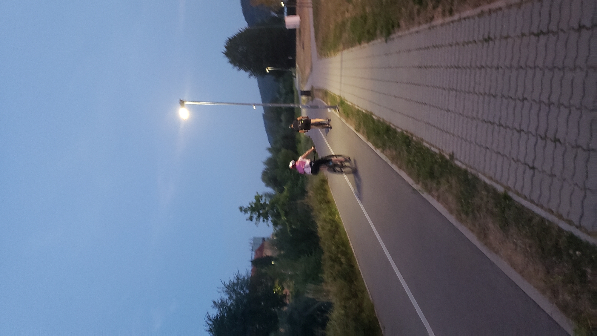 At dusk, two individuals are cycling on a dedicated bike path alongside a paved pedestrian walkway. The bike path, marked with a white line, is to the left of the pedestrian path. A single streetlamp illuminates the scene, casting a soft glow. The environment is characterized by trees and vegetation lining the path, with a building and a hillside visible in the background. The sky is a clear, pale blue, indicating the transition from day to night. This scene is captured in Prešov, Slovakia, likely a park or recreational area designed for outdoor activities.
