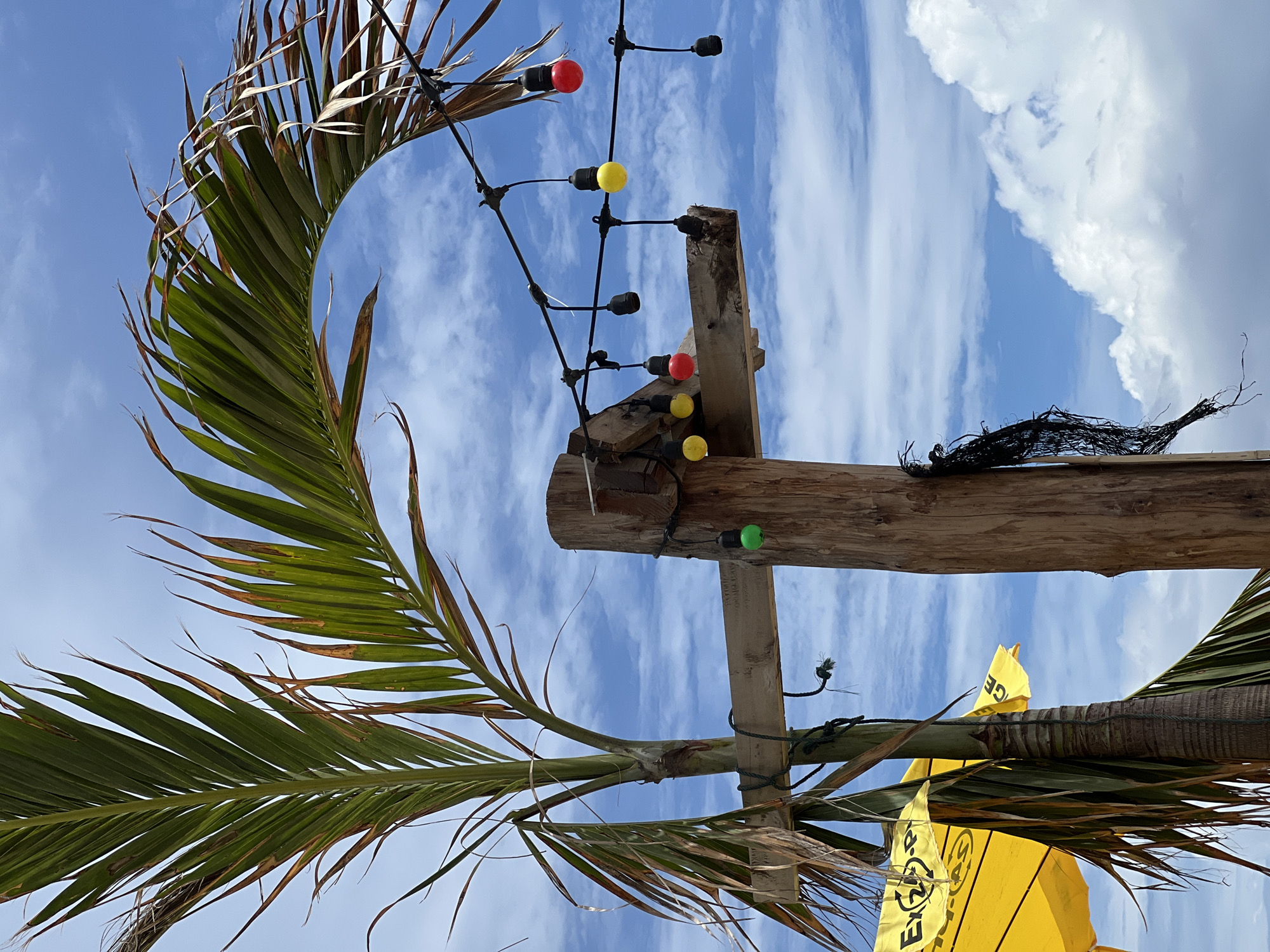 A close-up shot captures a weathered wooden post against a blue sky with scattered white clouds. A palm frond arches over the scene from the left, with some fronds appearing dry and brown. A string of colorful lights—red, yellow, and green—is strung across the frame, attached to the wooden post and a horizontal wooden beam. One green light bulb hangs from the beam. A dark, tattered net is also tied to the post. In the lower left, two yellow umbrellas are partially visible, with one displaying yellow lettering. The overall scene suggests an outdoor, possibly tropical, setting.