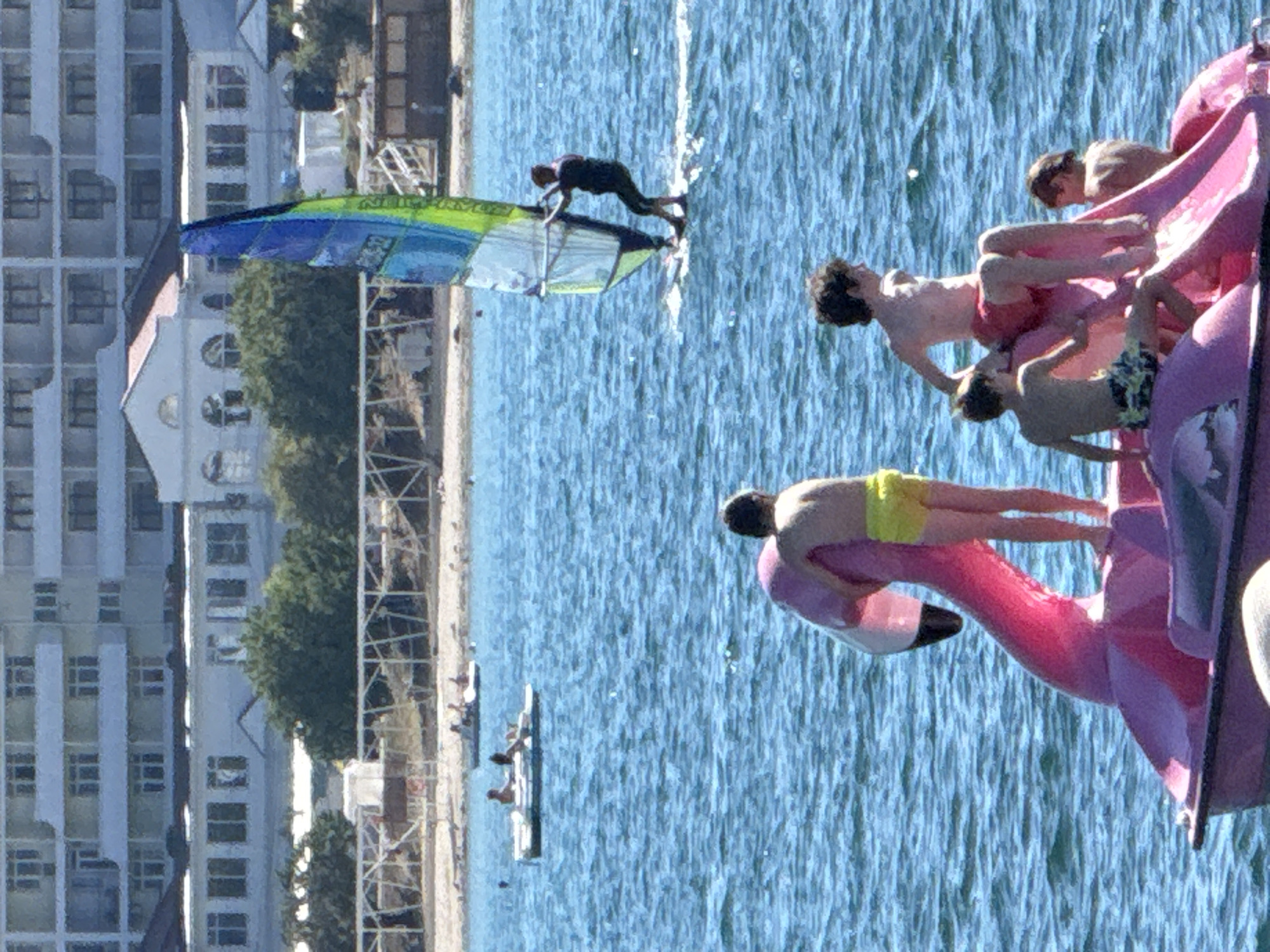 This is a vertical, medium-distance shot of a sunny day at a beach in Sevastopol, Ukraine, with several people engaged in various water activities. In the foreground, a pink flamingo-shaped paddleboat is partially visible, with three young boys sitting on it. One boy, wearing yellow swim trunks, stands at the front of the boat, facing away from the camera. Another boy, in camouflage swim trunks, is seated on the left side of the boat, facing towards the right. A third boy, wearing red swim trunks, is sitting on the right side of the boat, facing left. In the middle ground, a person is windsurfing on a blue and green sailboard, moving across the choppy blue water. Further back, two small pedal boats are visible with people in them. The background shows a sandy beach with trees and a multi-story building. The sky is clear and blue.