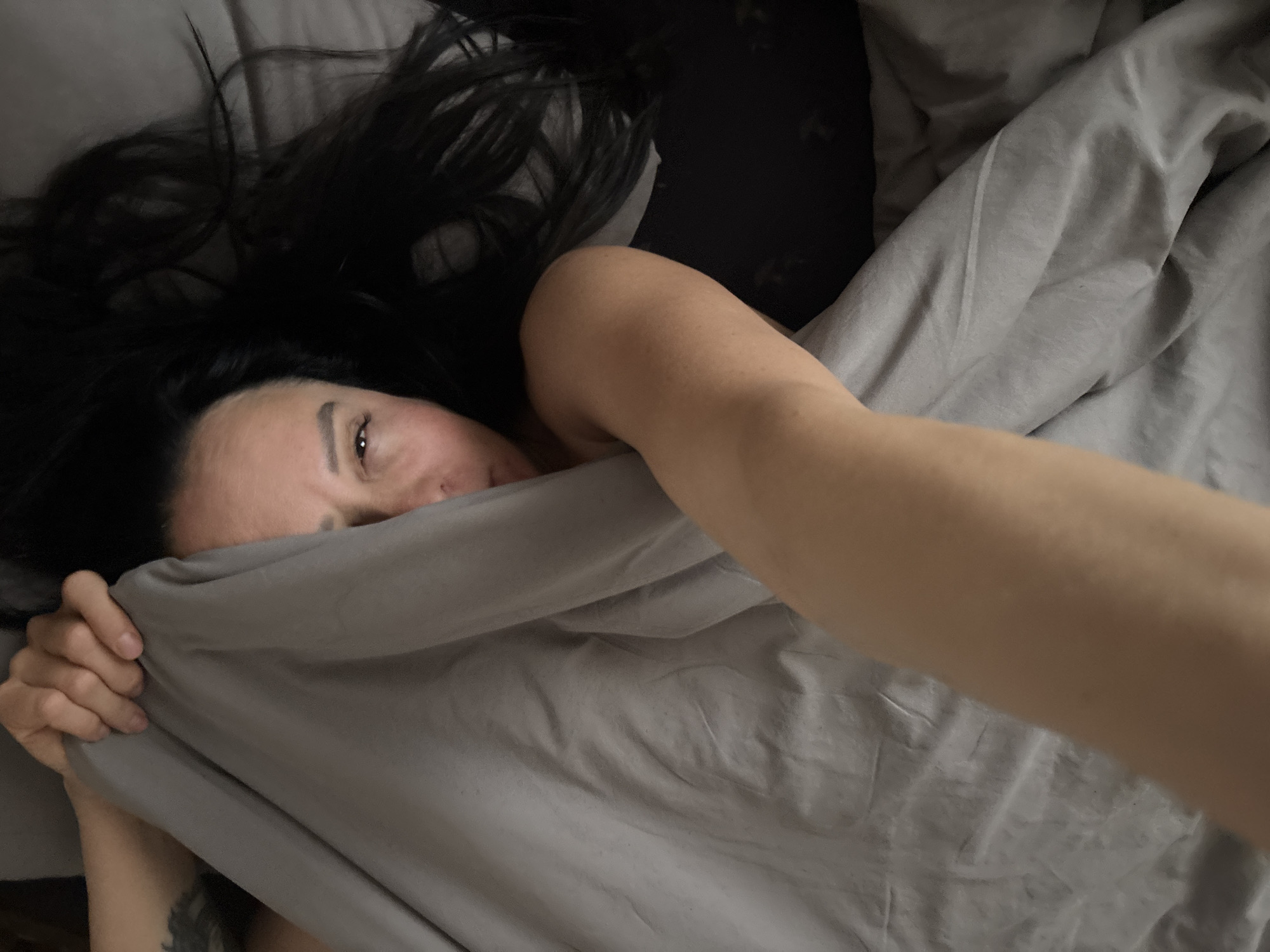 A woman with dark hair is lying in bed. She is mostly covered by a gray blanket, which she is holding up with her left hand, partially obscuring her face. Only her right eye is clearly visible. Her right arm is extended towards the camera. The bed has gray sheets and a dark gray headboard. The location is unknown.