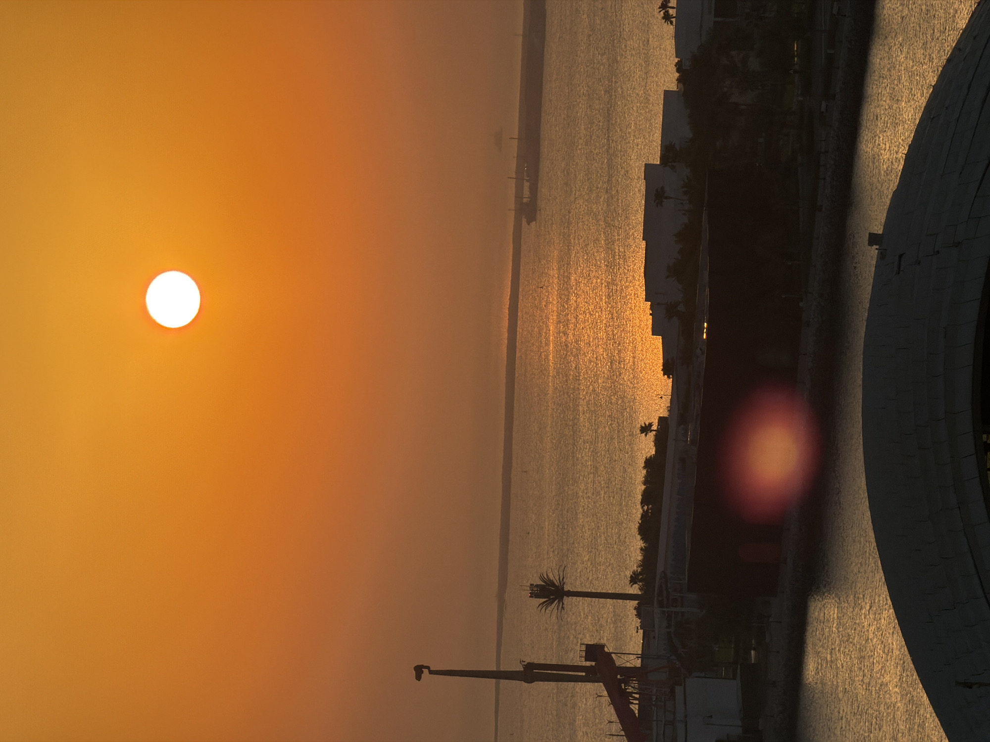 This is a high-angle, vertical, full shot of a sunset over a body of water in an urban setting. The sky is a gradient of orange and yellow, with a bright white sun positioned in the upper center. In the foreground, the curve of a dark, domed structure takes up the bottom quarter of the image. Behind this, a body of water reflects the sunset, with buildings and palm trees silhouetted along its edge. A large, dark building with a red door is prominent in the midground, with a blurry red and orange lens flare in front of it. To the left of this building, a tall structure with a crane-like extension is visible. In the distance, across the water, a long, low-lying structure that appears to be a port or industrial area is visible, with a large ship docked. The overall atmosphere is serene and captures the warm colors of a sunset.