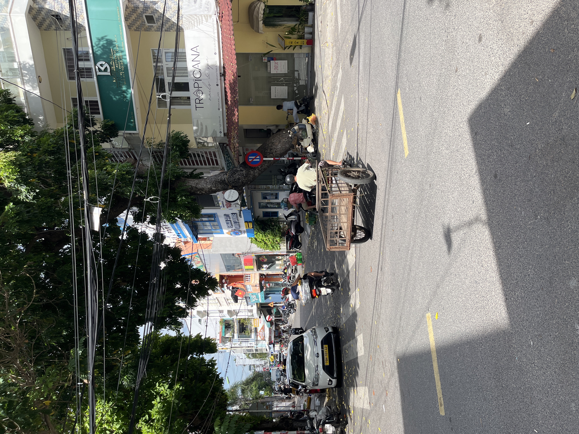 The image depicts a street scene in Nha Trang, Vietnam, on a sunny day. In the foreground, a three-wheeled vehicle with a trailer is being ridden across a pedestrian crossing. The vehicle is laden with goods. A white car with license plate "79H-101.42" is parked on the left side of the road.
The street is lined with multi-story buildings, some of which house businesses with visible signage. One building features a sign for "TROPICANA nails & spa." Numerous motorcycles are parked along the sidewalk and on the street. Overhead, a dense network of electrical wires crisscrosses the sky, partially obscuring the view of the buildings. Trees with dense green foliage overhang the street, casting shadows.
Several individuals are visible, mostly riding motorcycles or operating vehicles. The environment appears to be a busy urban commercial area, with typical street infrastructure like traffic lights and road markings. The overall impression is one of everyday life in a Vietnamese city.