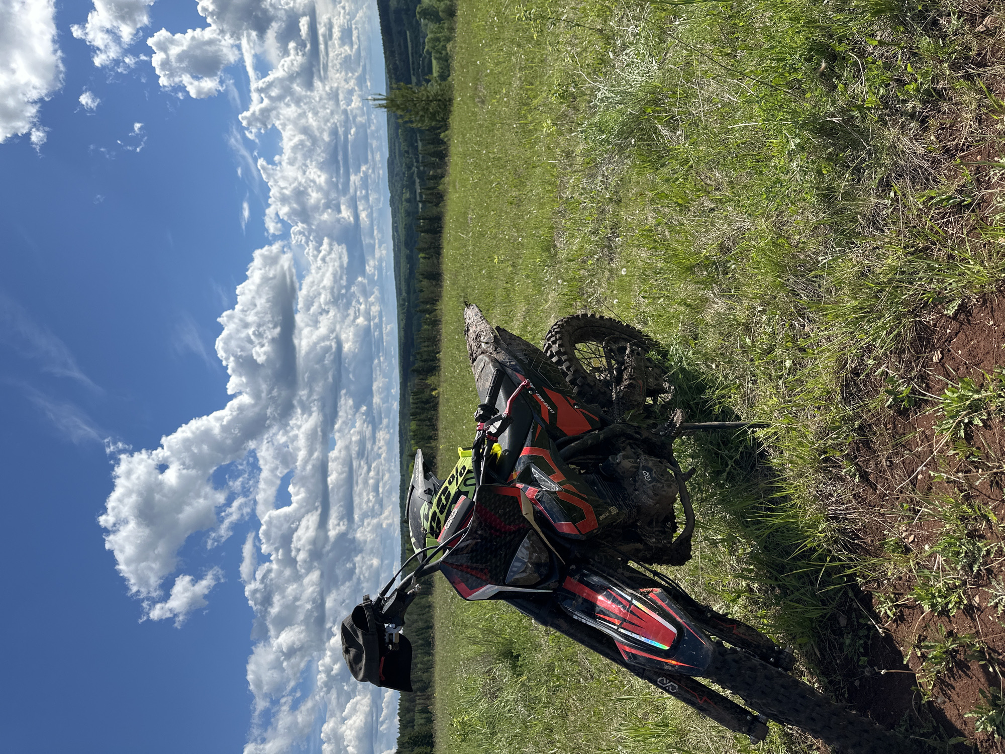 The image depicts a dirt bike lying on its side in a grassy field under a blue sky with fluffy white clouds. The dirt bike is black, red, and grey, covered in mud. A black cap hangs from the handlebars. The background includes a distant treeline. The location is Пепеляева, Russia.