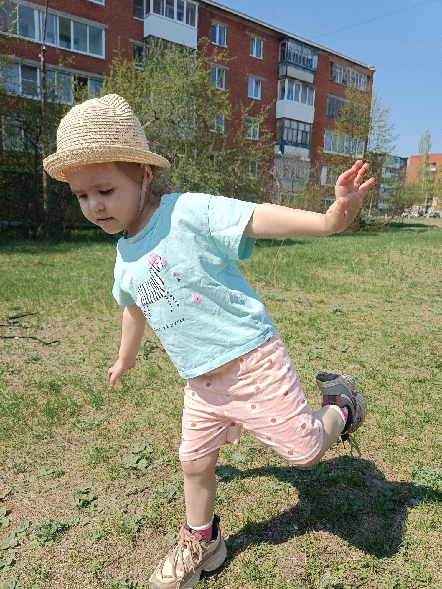 A young child stands in a grassy area in Noviy, Russia, attempting to balance on one leg. The child wears a straw hat, a light blue t-shirt with a zebra graphic, pink shorts with white polka dots, and brown sneakers. The child's right arm is extended to the side for balance, and the gaze is directed downwards. A multi-story brick apartment building with windows and balconies is visible in the background, along with green trees and clear blue sky.