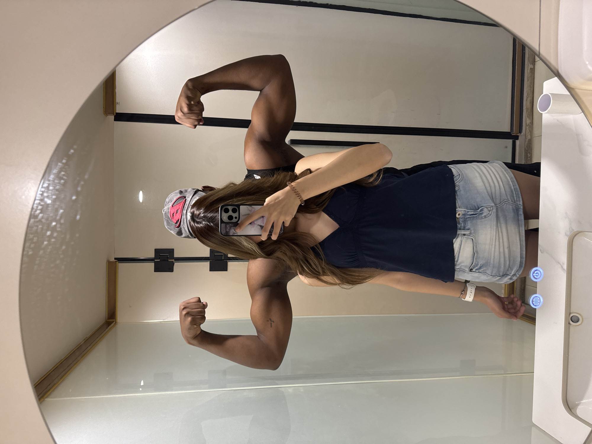 The image, rotated 90 degrees clockwise, captures a mirror selfie. The main subject is a young woman, partially obscured by her smartphone as she takes the photo. She has long brown hair and wears a dark blue top, light blue denim shorts, and a grey cap with a red design. A white watch or bracelet is visible on her left wrist.
Prominently featured in the reflection alongside her, or as a deliberate part of the scene, are the flexing bicep muscles of a person with dark skin. This individual's arms are muscular, and one arm displays a small cross tattoo. They appear to be wearing dark attire.
The setting is an indoor space, likely a modern bathroom, identifiable by the large, rounded-top mirror and illuminated touch controls on a white surface to the right. These blue-lit buttons display a power symbol and a defogger/heating icon, suggesting a smart mirror or vanity unit. The background reflected in the mirror includes light-colored walls and dark horizontal frames, possibly part of a shower enclosure or shelving. The activity is a playful mirror selfie, creating a humorous contrast between the two reflected figures. Time of day and weather are not discernible, and no distinct text is legible apart from the universal icons.