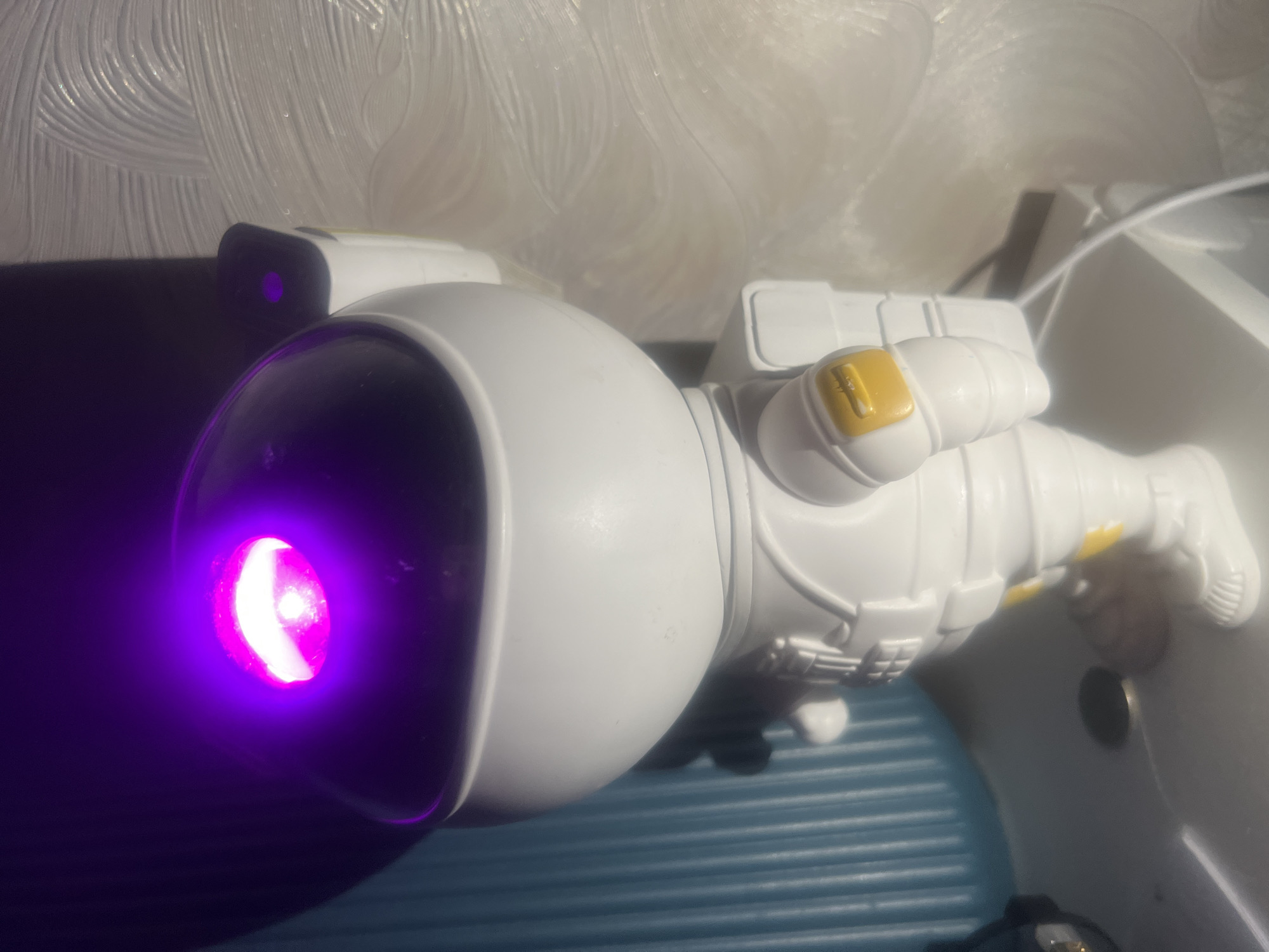 A close-up shot presents a white astronaut figurine with a purple light emanating from its helmet. The figurine is resting on a textured blue surface. The background includes a white wall with a textured, swirled pattern and a white object with a cable. The location is unknown, and no information suggests it is in Chaikovsky, Russia.