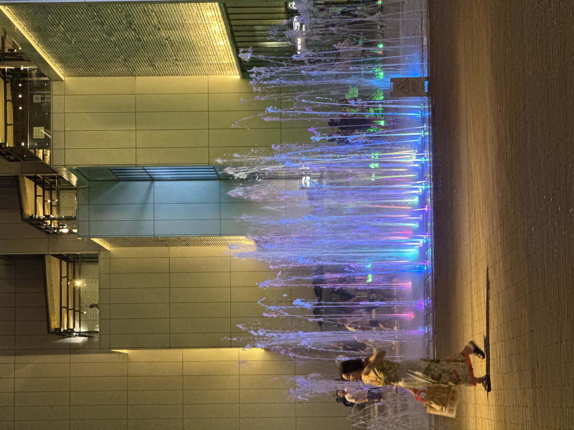 A nighttime exterior scene shows a woman walking on a paved walkway in front of a brightly lit fountain with multicolored jets of water. The woman is wearing a long, patterned dress and carrying a shopping bag. Several other people are visible in the background, near the fountain. The fountain is located in front of a modern building with illuminated sections and a patterned facade. The scene is likely in a public urban space.