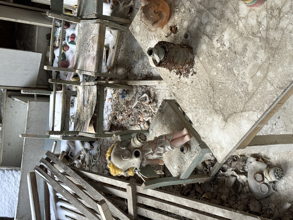 This image captures an interior scene in the city of Prypiat, Ukraine, consistent with an abandoned classroom or playroom. No human subjects are present. In the mid-ground, a plastic doll with yellow synthetic hair, wearing a light-colored dress, sits on a distressed light-green wooden chair. A grey gas mask with two eye lenses and a cylindrical filter covers the doll's head. To the right, a dust-laden light-colored wooden table supports a heavily rusted cylindrical object and a partially visible orange, possibly plastic, item. On the floor to the lower left, a flattened grey gas mask lies amidst debris. The background reveals additional small, similarly distressed wooden chairs and a floor strewn with debris, dust, scattered small, colorful toys, and patches of snow. To the far left, a pile of wooden planks is visible. The environment exhibits significant decay, dust accumulation, and signs of prolonged abandonment and exposure to cold elements.