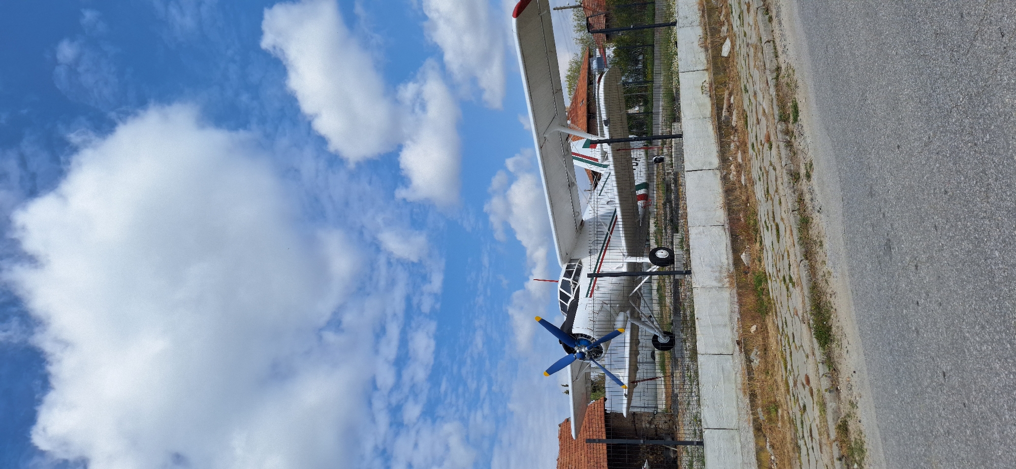 A white biplane with blue propellers is parked in Kapatovo, Bulgaria. The plane is positioned next to a low fence and a concrete curb, with a paved road running adjacent to the curb. The sky, filled with puffy white clouds against a blue backdrop, dominates the upper portion of the frame. The plane has red and green markings on its tail. Red brick buildings and greenery are visible behind the fence.