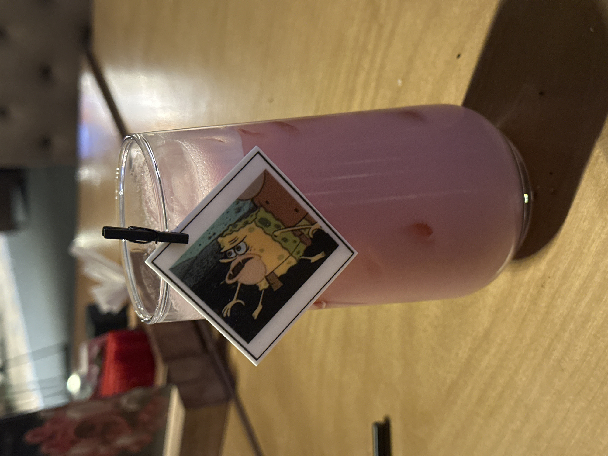 The image shows a pink beverage in a clear glass, oriented horizontally on a wooden table. A square sticker depicting Spongebob Squarepants is clipped to the rim of the glass. The table has a light brown color. In the blurred background, a portion of an interior setting is visible, suggesting a restaurant or bar with red and white elements. There is no indication of Izhevsk, Russia or any other specific location.