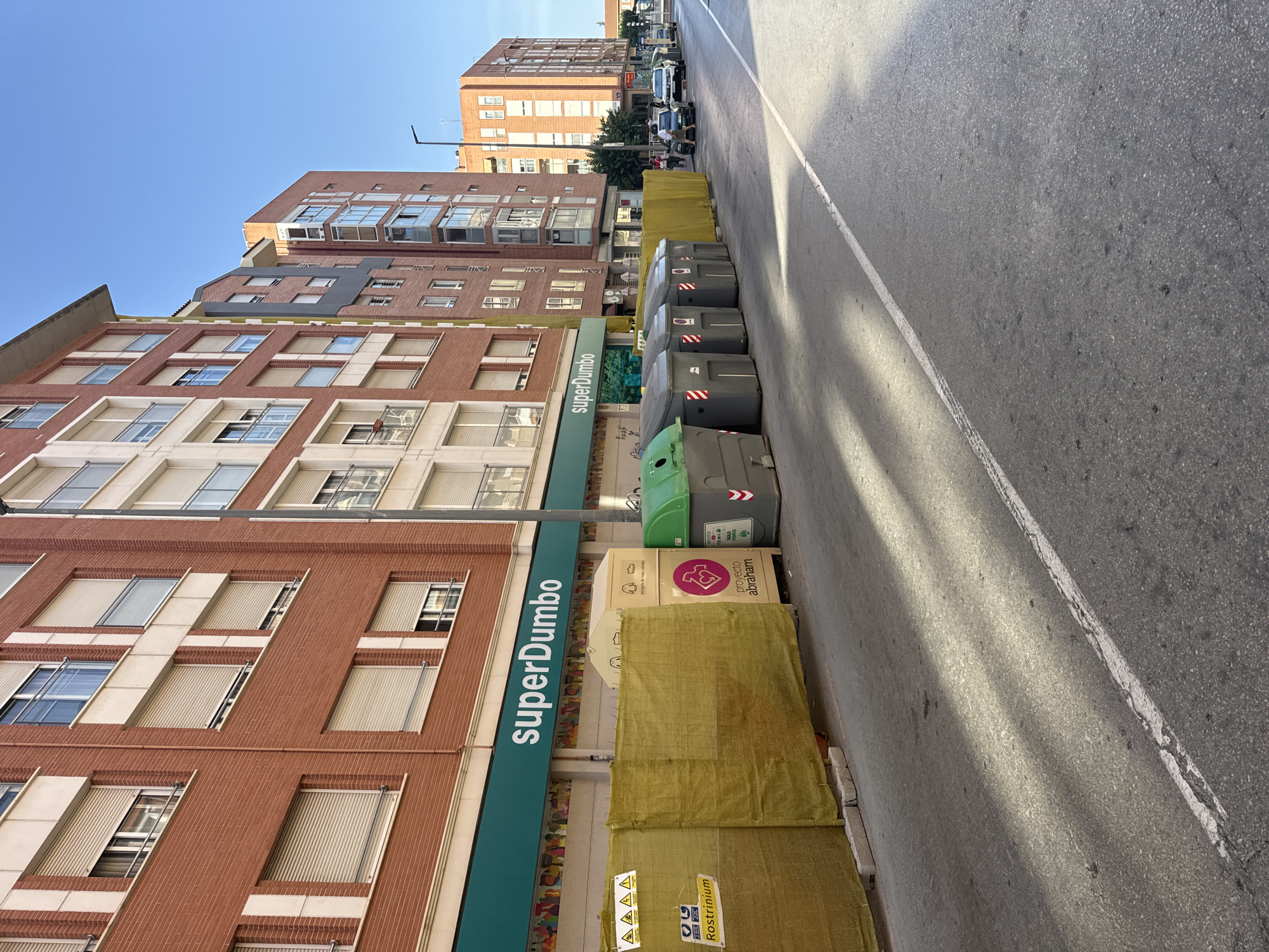 This image depicts a street scene in the city of Murcia, Spain, likely during daylight hours with clear skies. The foreground shows an asphalt road with a white line marking the edge of a lane. Sunlight casts distinct shadows across the road, suggesting a mid-morning or late afternoon time. Along the right side of the road, a row of grey and green refuse bins is visible. Behind the bins, a yellow construction barrier or screen runs along the sidewalk.
The buildings lining the street are modern apartment complexes with brick facades and white architectural accents. The lower floors of these buildings house commercial establishments, one of which has a large teal sign with white lettering that reads "superDumbo". Above the sign, the buildings feature numerous windows, many of which are covered by beige roll-down shutters.
Further down the street, other buildings of varying heights and architectural styles are visible. Several cars are parked or driving on the street, and a few individuals can be seen walking or standing near the businesses. The overall impression is that of a typical urban environment undergoing some form of construction or renovation, indicated by the barriers and the presence of waste bins.