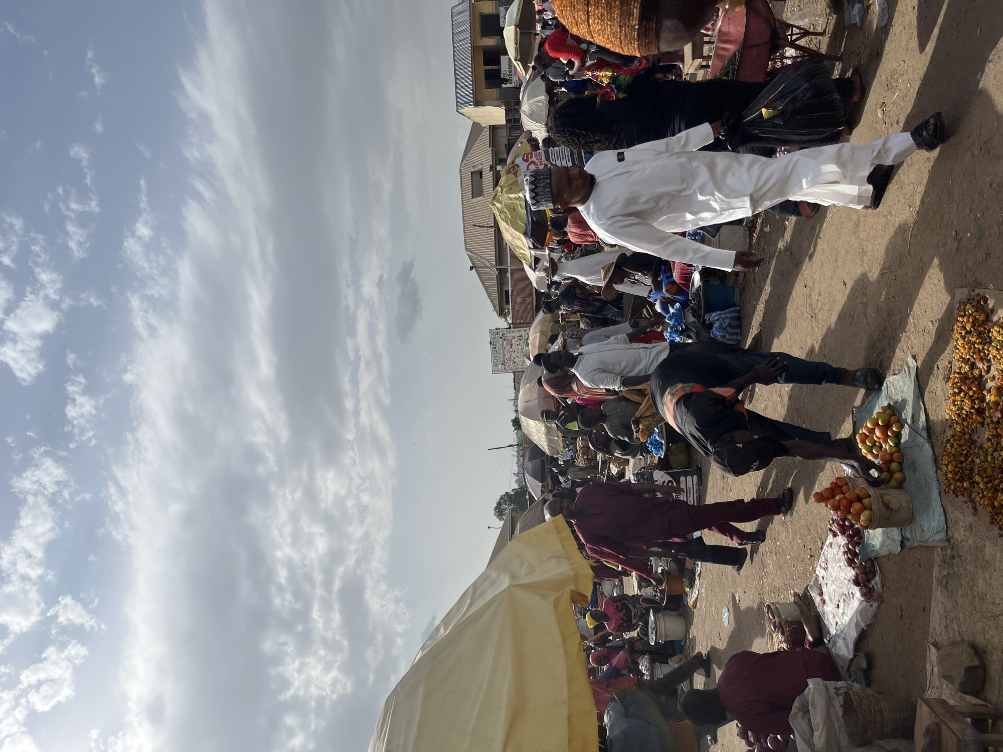 This image depicts a bustling outdoor market scene in Rafin Par, Nigeria, under a cloudy sky. Numerous individuals are engaged in various activities, suggesting a vibrant commercial atmosphere. In the foreground, a man in a white traditional outfit walks towards the right, carrying a black plastic bag. To his left, another person in a black shirt and jeans bends over, possibly attending to goods on the ground.
Scattered throughout the market are stalls with various items for sale. A prominent yellow umbrella shades a section of the market. In the middle ground, several individuals are seen interacting, some appearing to be vendors and others customers. Various goods, including produce such as tomatoes and what appear to be onions, are displayed on the ground and in containers. Umbrellas of different colors and sizes dot the landscape, providing shade.
In the background, buildings with pitched roofs are visible, indicating a semi-urban or rural setting. The ground is covered in dirt and dust, typical of an outdoor market. The overall impression is one of lively economic activity.