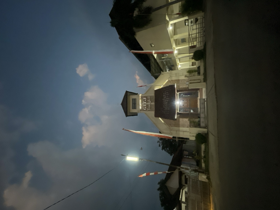 A nighttime street-level view in Trangkil, Indonesia, displays the illuminated exterior of the GITJ Trangkil building. The central, cream-colored structure features a gabled facade, a prominent entrance with dark double doors under a decorative awning, and a belfry-like tower containing a window at its apex. Warm artificial lights highlight the building's architectural details and the "GITJ Trangkil" signage. Two Indonesian national flags, red and white, are visible on poles flanking the main building. To the left, a bright streetlamp stands beside overhead power lines and a residential building partially obscured by a fence. To the right, an adjacent, illuminated multi-story building is partially visible, with a palm tree in the far right. A dark, paved road occupies the immediate foreground. The sky above is a dark blue-grey with scattered white clouds. No people are present in the scene.