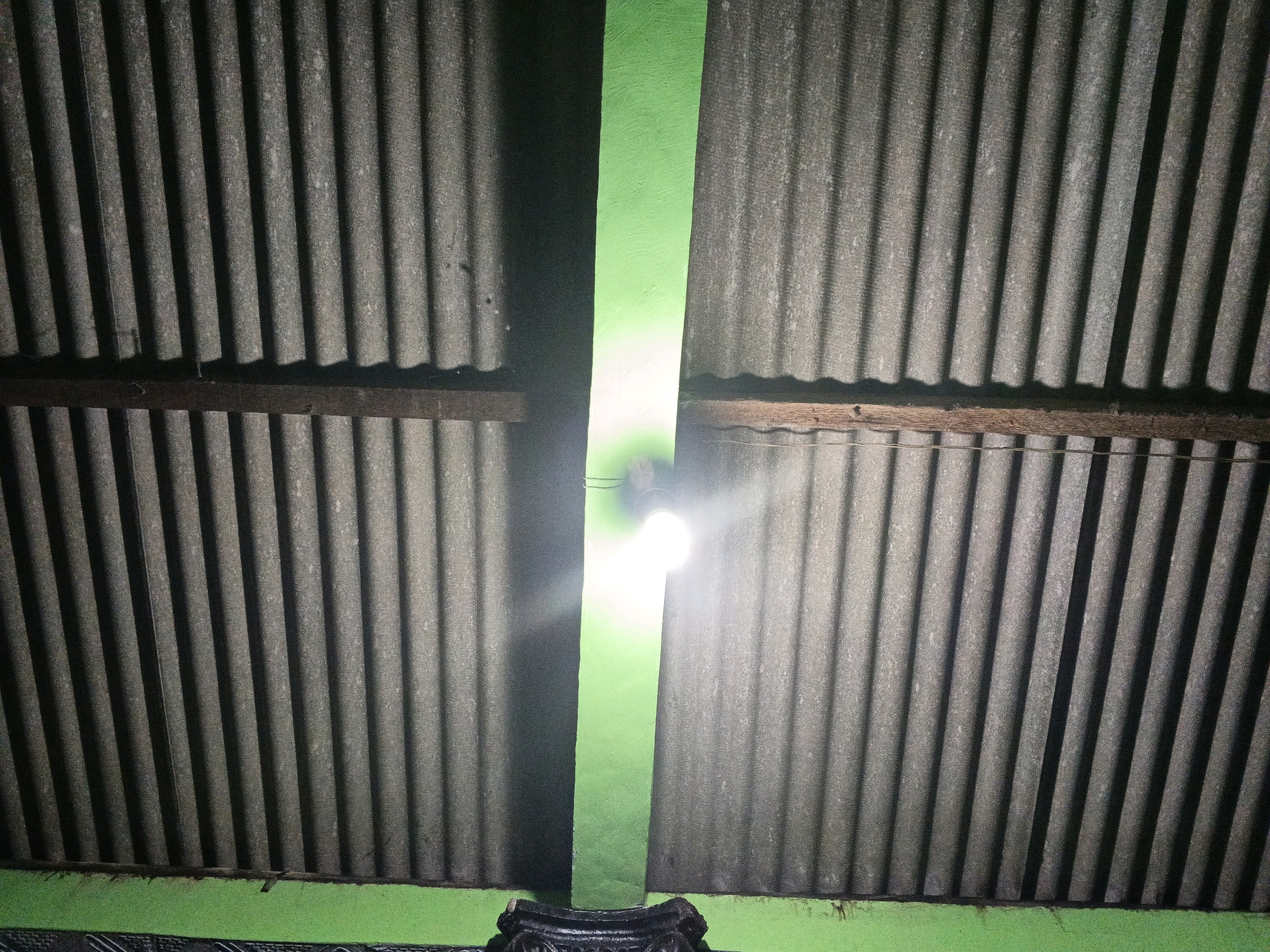 The image shows a view looking upwards at a corrugated metal roof, with a bright green wall running vertically between two sections of the roof. A lightbulb is visible in the gap between the roof sections, illuminating the green wall and casting a bright glare. Wooden beams support the roof. The lower part of the image shows a green painted surface, possibly a wall or the edge of a structure, with a dark, ornate object at the bottom. The scene appears to be an outdoor or semi-outdoor area. The overall impression is one of simple, utilitarian construction, possibly in a residential or rural setting. Without further context, it is not possible to determine the specific location in Wates, Indonesia, but the materials and lighting suggest a common building style.