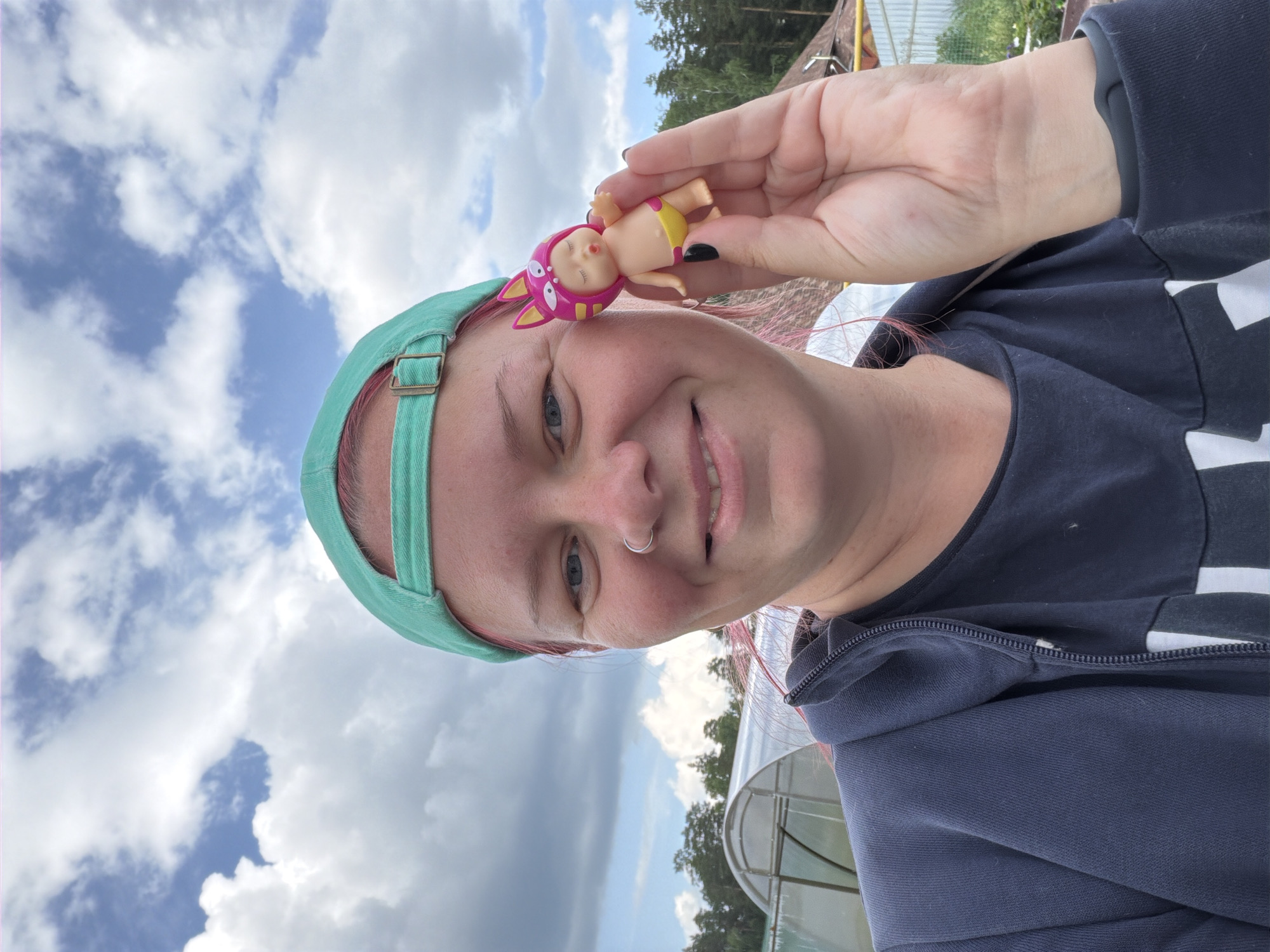 A medium shot captures a woman with pink-streaked hair wearing a teal baseball cap and a dark blue hooded jacket. She is holding up a small, doll-like figurine with a pink animal-eared hat and yellow bottoms in her right hand, displaying it to the camera. Her left hand is out of frame. The woman is smiling and looking at the camera, with a silver nose ring visible. In the background, a greenhouse-like structure with a curved translucent roof is situated in a rural or semi-rural outdoor setting, with trees and a cloudy blue sky visible. This location is consistent with the outskirts of Chaikovsky, Russia.