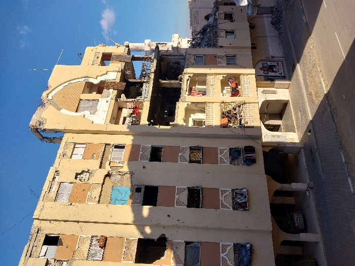 The image displays a multi-story building in Khan Yunis, Palestinian Territory, exhibiting severe structural damage under a clear blue sky with sparse clouds. The building, constructed with beige and reddish-brown sections, features at least six visible floors. Extensive damage is concentrated on the upper floors and the right side, where the roof and upper sections are largely demolished, exposing interior rooms, concrete debris, and twisted rebar. A decorative arched element on the upper-right facade is partially intact. Multiple window openings across the entire facade are blown out, some blackened by fire, while a bright blue tarp covers an opening on the fourth floor from the bottom. Numerous smaller impact marks are visible across the building's surface. Several balconies on the mid-levels show damaged railings and debris, yet clothes are hanging on lines and from window frames on these and lower floors, suggesting recent human presence or activity. The ground floor features arched open spaces, one of which houses a parked silver sedan. A paved street runs in front of the building, displaying minor cracks and scattered debris, with a significant pile of rubble accumulated against the building's right side at street level. A partially visible storefront sign with red and white graphics is on the ground floor on the right. No people are visible on the street or in the immediate vicinity of the building.