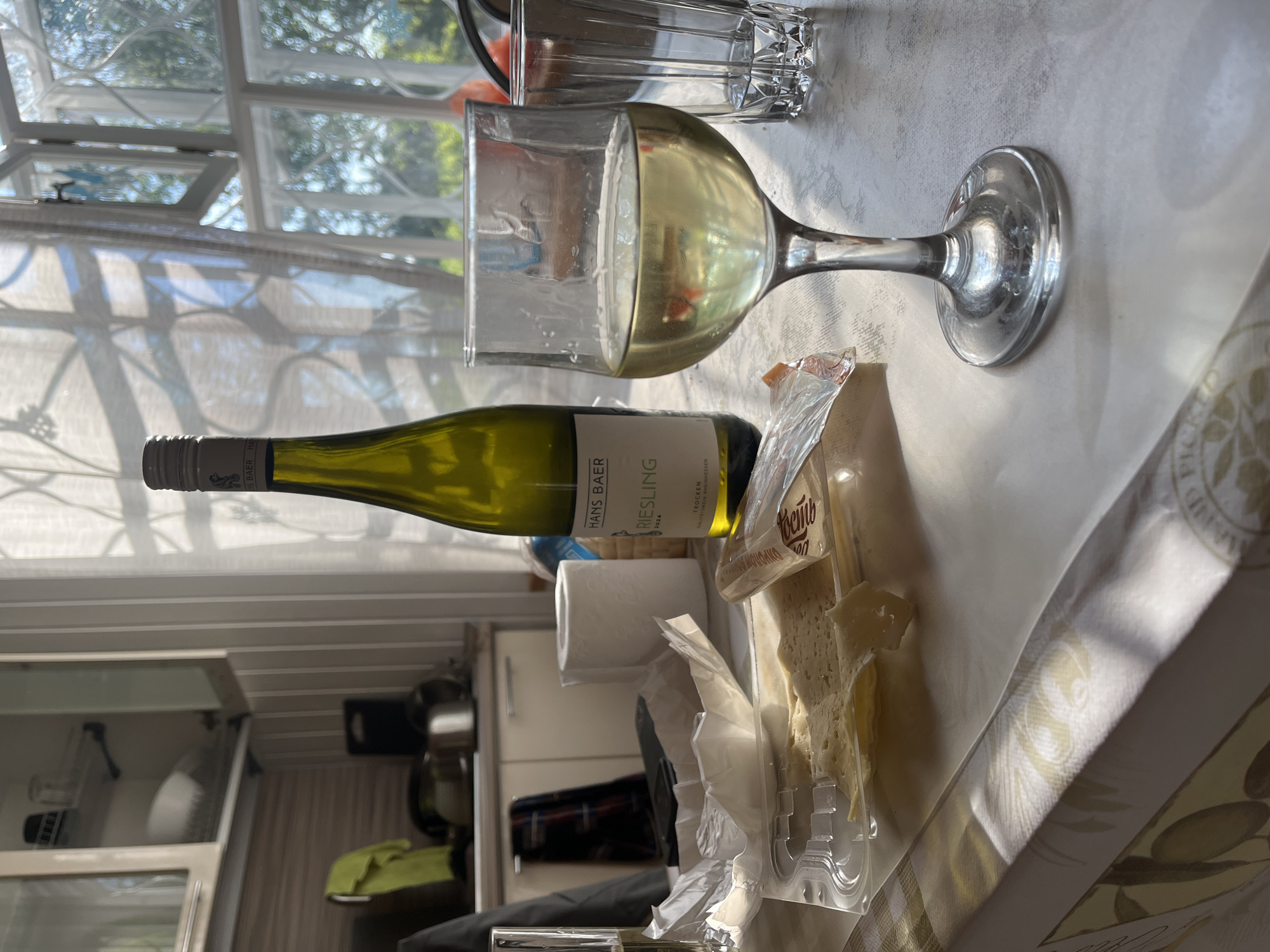 The image shows a tabletop scene in an indoor setting. A bottle of white wine, labeled "Hans Baer Riesling", lies horizontally. A wine glass containing white wine is positioned near the bottle. Next to the wine and glass, there is a package of sliced cheese on a plastic tray. A roll of toilet paper is in the background. The backdrop includes a window with sunlight filtering through curtains and a glimpse into what appears to be a kitchen with cabinets and utensils. The location in Пятница, Russia, cannot be confirmed from the image.
