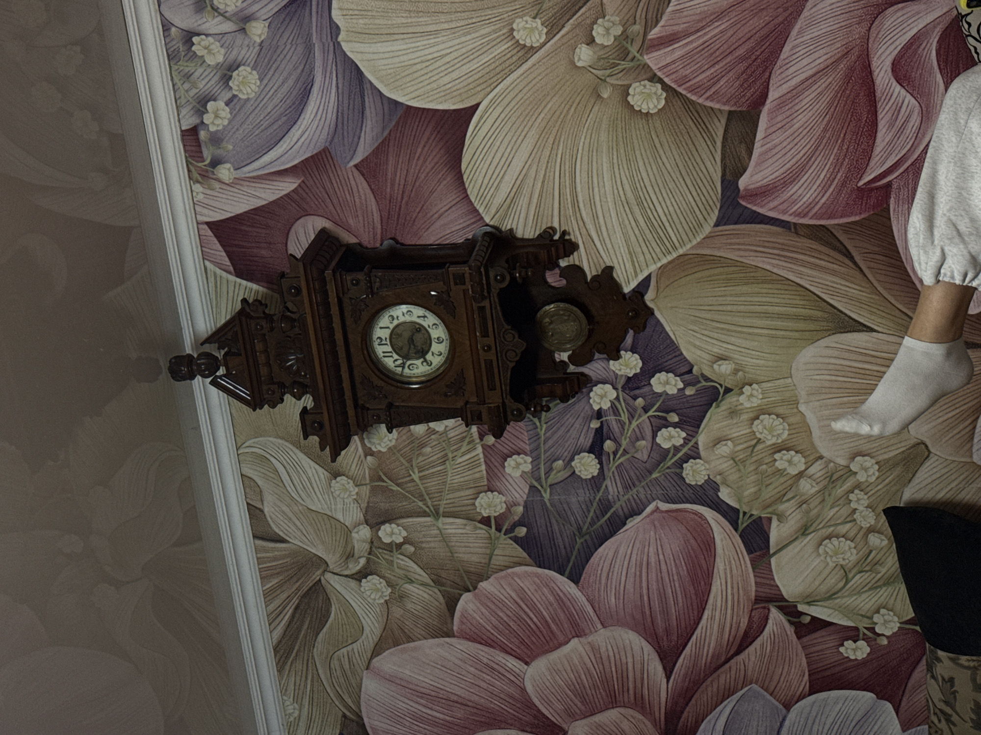 A medium, eye-level shot captures an ornate, antique, wooden cuckoo clock hanging on a wall adorned with floral wallpaper. The clock features Roman numerals, decorative carvings, and a pendulum. A person, clad in grey clothing and white socks, sits to the right of the clock. A portion of a black object is visible at the bottom right of the frame. The wallpaper design consists of large, stylized flowers in shades of white, purple, and pink. The scene is situated in an indoor setting in an unspecified city.