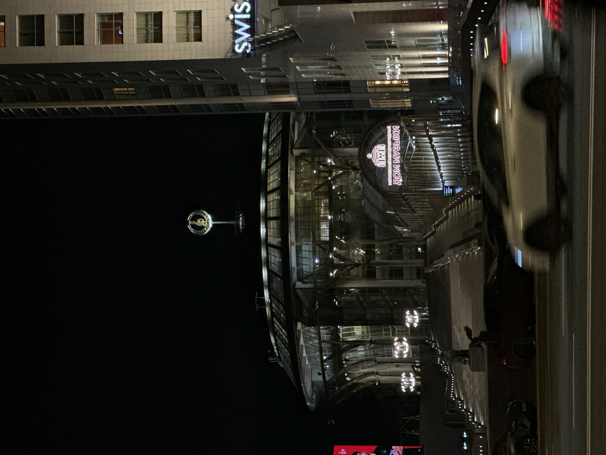 Here is a description of the image:
The image captures an urban scene at night. Prominently displayed is the Moscow International House of Music, identified by its illuminated Russian signage. Adjacent to it stands a high-rise building with "swiss" displayed vertically, indicating a Swissôtel. A musical note sculpture on a tall stand is visible in the dark space. A street runs next to the building, with cars captured in motion. Several parked vehicles are also seen. The lighting from buildings and streetlights creates a mixture of bright and dark areas, emphasizing the nocturnal setting.