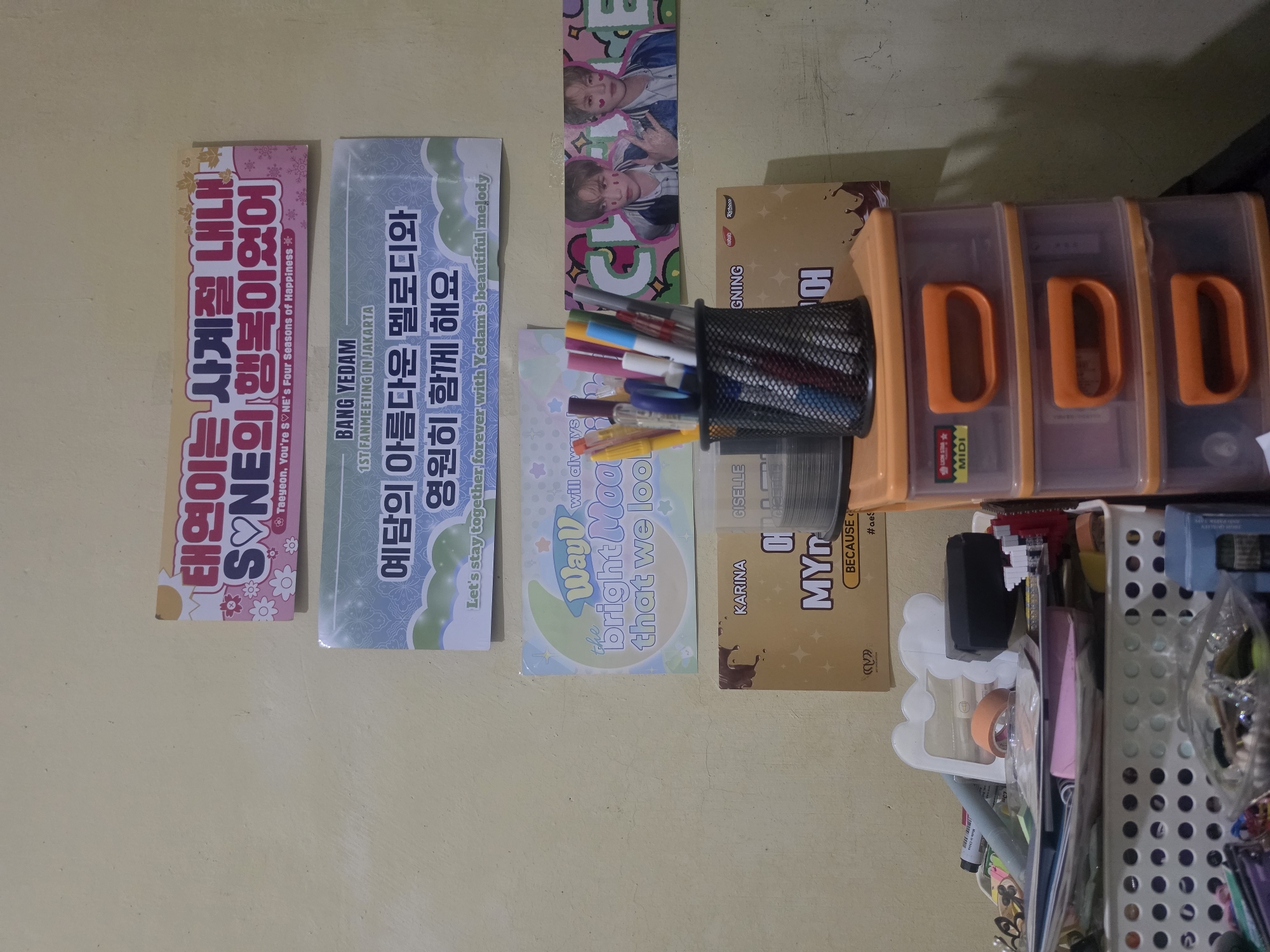 The image shows a collection of items placed on a light yellow surface. There are several banners with Korean text and English translations. The banners feature names such as "Taeyeon," "BANG YEDAM," and "WayV." A container filled with pens and markers sits next to these banners, and a box of "Richoco" snacks is also present. Adjacent to these items are three orange and clear plastic storage containers labeled "MIDI," and a white, perforated basket is overflowing with miscellaneous items, including office supplies, tape, and other unidentified objects. The scene is cluttered and suggests a personal or organizational space.